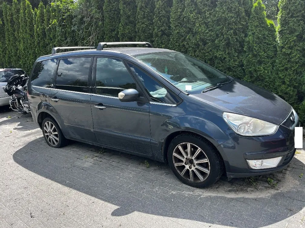 Ciemnoszary samochód typu minivan zaparkowany na kostce brukowej na tle wysokiego, zielonego żywopłotu. Pojazd posiada relingi dachowe oraz wieloramienne felgi aluminiowe. W tle po lewej stronie widoczny jest fragment innego samochodu oraz motocykl.