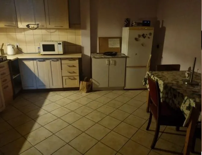 Kuchnia z jasnymi szafkami wiszącymi i dolnymi, mikrofalówką na blacie, białą lodówką z magnesami, małym szafkowym stolikiem z czarną patelnią, stołem z kwiecistym obrusem i krzesłami. Na podłodze z kafelkami leży brązowa torba papierowa. Oświetlenie jest przygaszone, a na ścianie widoczna jest tablica z niewyraźnym tekstem.