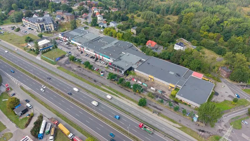 Zdjęcie lotnicze przedstawiające kompleks handlowo-usługowy z dużymi, płaskimi dachami budynków, otoczony terenami zielonymi i lasem. Obok kompleksu przebiega wielopasmowa droga szybkiego ruchu z widocznymi samochodami i ciężarówkami. W tle widoczne są mniejsze budynki mieszkalne oraz biurowiec o nowoczesnej architekturze.