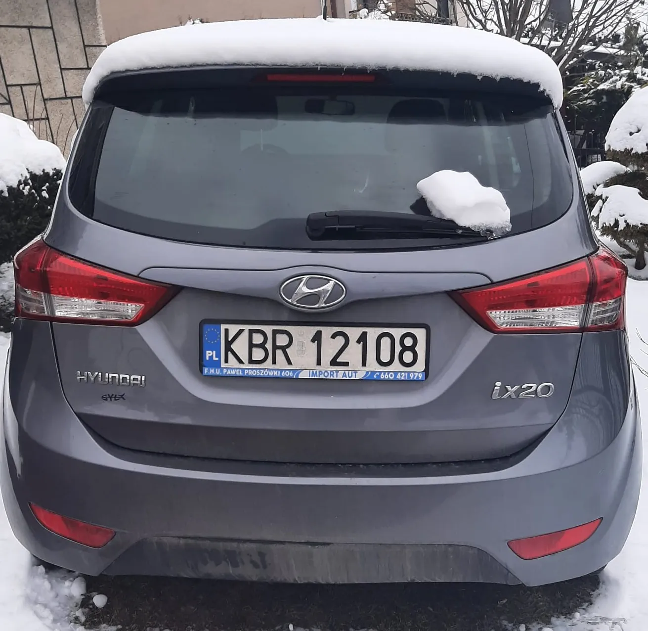 Tył szarego samochodu Hyundai ix20 z tablicą rejestracyjną KBR 12108 (PL), pokryty śniegiem na dachu i szybie. Widoczne logo marki, napisy "HYUNDAI" i "ix20". Tło: zasneżone krzewy i fragment budynku.