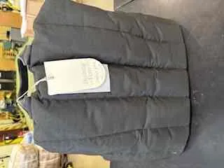 Czarna pikowana kamizelka leżąca na ciemnej powierzchni. Na kamizelce widoczny biały etykietka z nieczytelnym tekstem. W tle rozmyty widok wnętrza sklepu z półkami i przedmiotami, w tym butelką wody po lewej stronie.