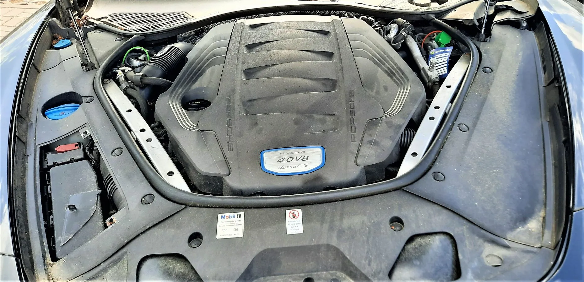 Widok z góry na komorę silnika samochodu marki Porsche. Centralnie znajduje się czarna osłona silnika z napisem 'PORSCHE' po obu stronach oraz niebieską tabliczką z napisem '4.0 V8 diesel S'. Wokół silnika widoczne są elementy mechaniczne, przewody oraz plastikowe osłony nadwozia. Na dolnej części obrazu widoczne są naklejki informacyjne, w tym jedna marki Mobil 1.