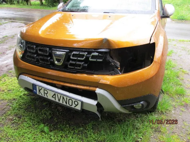 Przednia część uszkodzonego pomarańczowego samochodu marki Dacia. Widoczne uszkodzenia pasa przedniego po prawej stronie (z perspektywy widza), w tym pęknięty zderzak i uszkodzona atrapa chłodnicy. Tablica rejestracyjna z napisem KR 4VN09 oraz oznaczeniem PL. W tle mokra droga, trawa i drzewa. W prawym dolnym rogu obrazu widoczna data 11/06/2023.