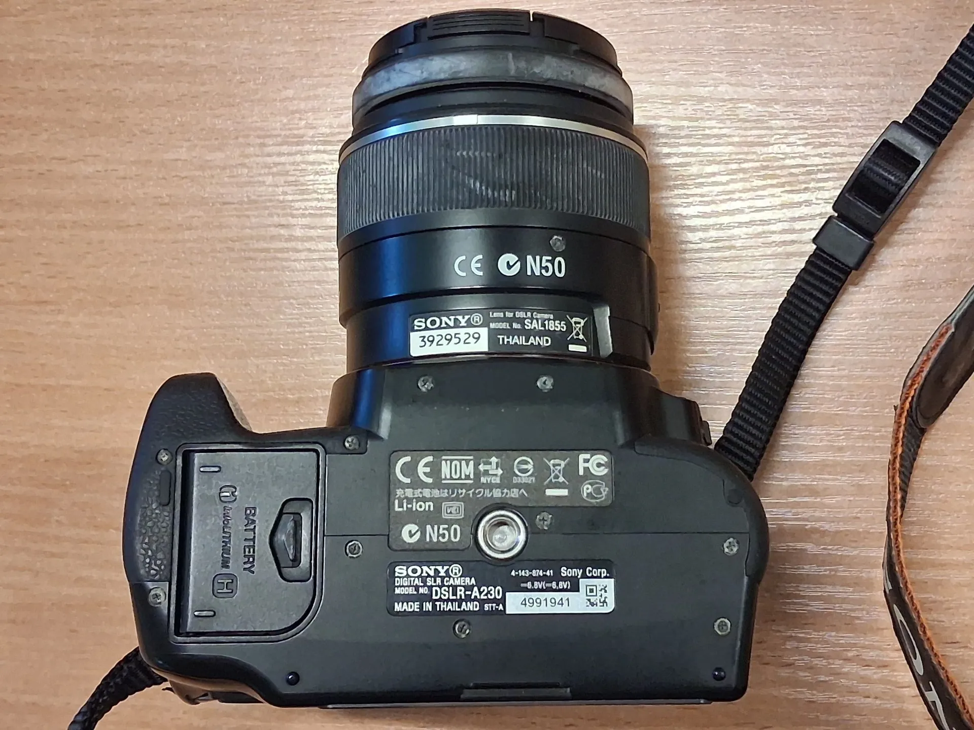 Czarny aparat cyfrowy Sony z obiektywem, leżący na drewnianym blacie. Widoczny jest spód urządzenia z naklejkami informacyjnymi. Na obiektywie znajduje się napis 'SONY Lens for DSLR Camera MODEL No. SAL1855 3929529 THAILAND' oraz oznaczenie 'N50'. Na spodzie korpusu widnieje napis 'SONY DIGITAL SLR CAMERA MODEL No. DSLR-A230 MADE IN THAILAND 4991941' oraz oznaczenia techniczne, w tym 'Li-ion' i 'N50'. Po prawej stronie widoczny jest czarny pasek do przenoszenia aparatu.