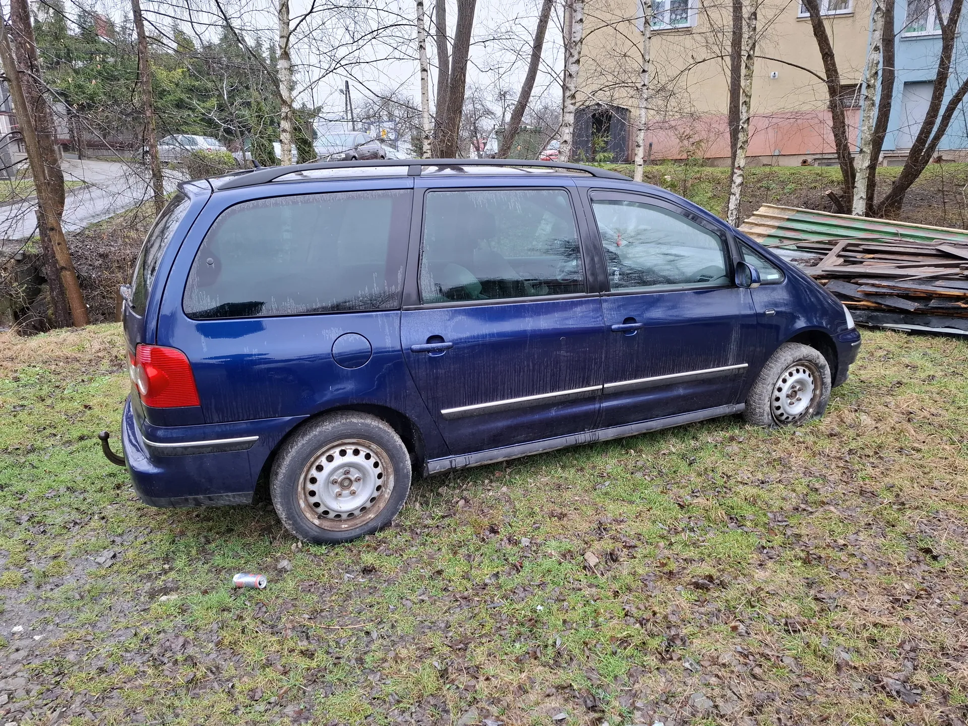 Niebieski minivan zaparkowany na trawie z suchymi liśćmi. Pojazd ma zabrudzoną karoserię, metalowe felgi i czerwone światło tylne. W tle widoczne są drzewa bez liści, budynki mieszkalne (żółty i niebieski), stos desek oraz pusta puszka na ziemi. Trawa jest częściowo zasłana suchymi liśćmi, a na tle droga i inne samochody.