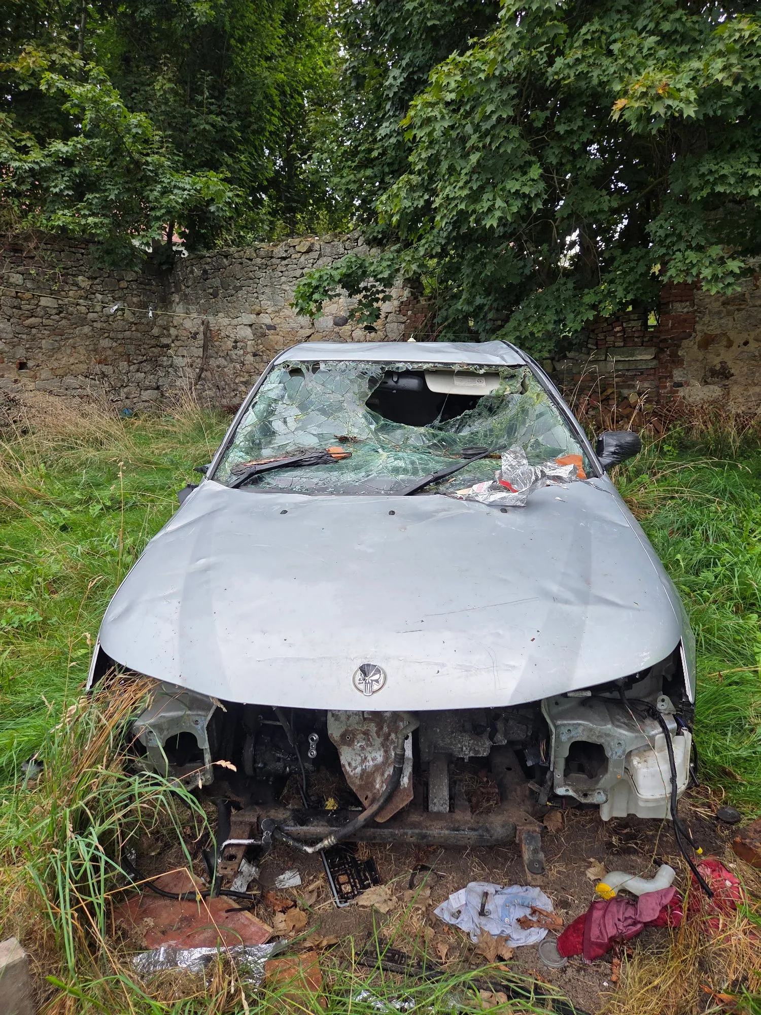 Zdjęcie zniszczonego, srebrnego samochodu osobowej marki Alfa Romeo, który stoi na trawiastym terenie przed kamiennym murem i drzewami. Pojazd jest poważnie uszkodzony: przednia część nadwozia jest całkowicie zdemolowana, brakuje zderzaka i reflektorów, a przednia szyba jest całkowicie rozbita. Wewnątrz samochodu i wokół niego widoczne są fragmenty gruzu i śmieci.