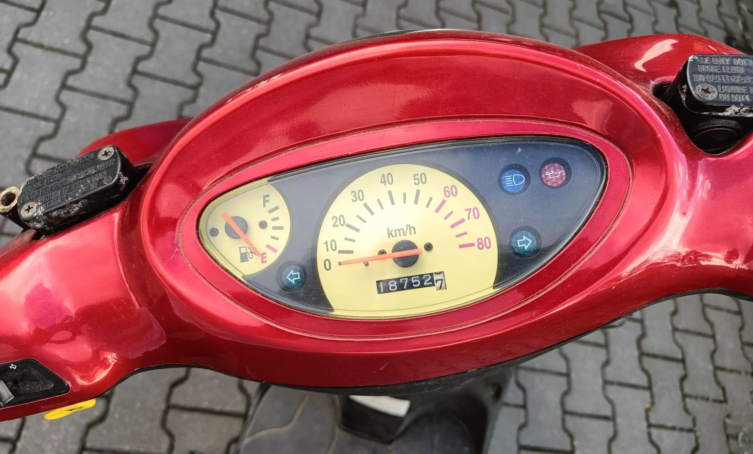 Zbliżenie na czerwony kokpit skutera. W centrum znajduje się żółty licznik prędkości z analogową wskazówką, skalą do 80 km/h i oznaczeniem 'km/h'. Poniżej znajduje się licznik przebiegu wskazujący wartość 18752. Po lewej stronie widoczny jest wskaźnik poziomu paliwa (skala F-E), a po prawej trzy kontrolki: niebieska, czerwona i zielona ze strzałką kierunkową. Obudowa skutera jest w kolorze intensywnej czerwieni.
