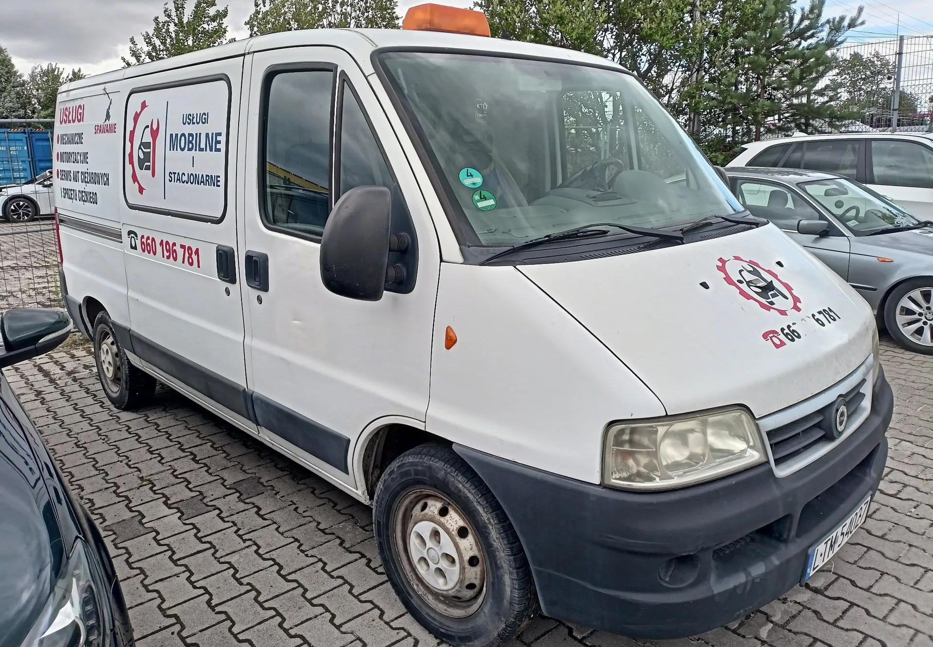 Biały samochód dostawczy zaparkowany na kostce brukowej. Na boku pojazdu znajduje się napis: 'USŁUGI SPRAWANIE MECHANICZNE, ELEKTRYCZNE, DIAGNOSTYKA, SERWIS AUT DOJEZDNYCH, SPRZĘTU CIĘŻKIEGO', a obok logo z kluczem i napisem 'USŁUGI MOBILNE STACJONARNE' oraz numerem telefonu '660 196 781'. Podobne logo i numer telefonu znajdują się na masce samochodu. W tle widoczne są inne zaparkowane samochody oraz drzewa.