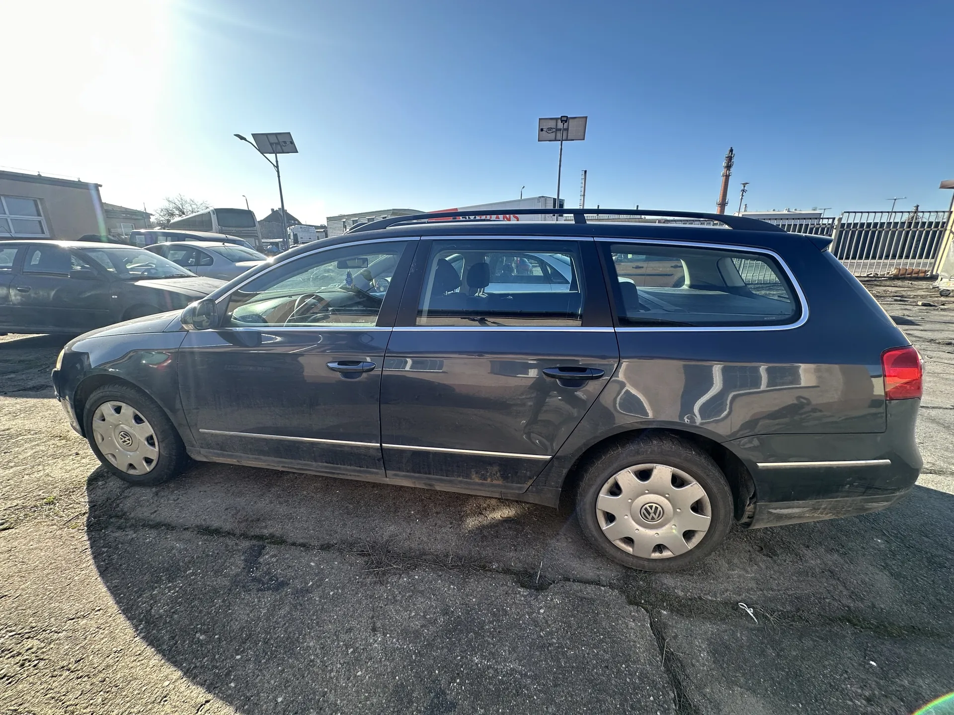 Ciemnoszary samochód osobowy typu kombi marki Volkswagen z białymi listwami nad kółami i dachowym bagażnikiem. Pojazd stoi na popękanej nawierzchni asfaltowej. W tle widoczne są inne samochody, budynki, latarnie z panelami słonecznymi oraz jasne niebo bez chmur. Słońce świeci silnie, tworząc odblask na lewej stronie obrazu.