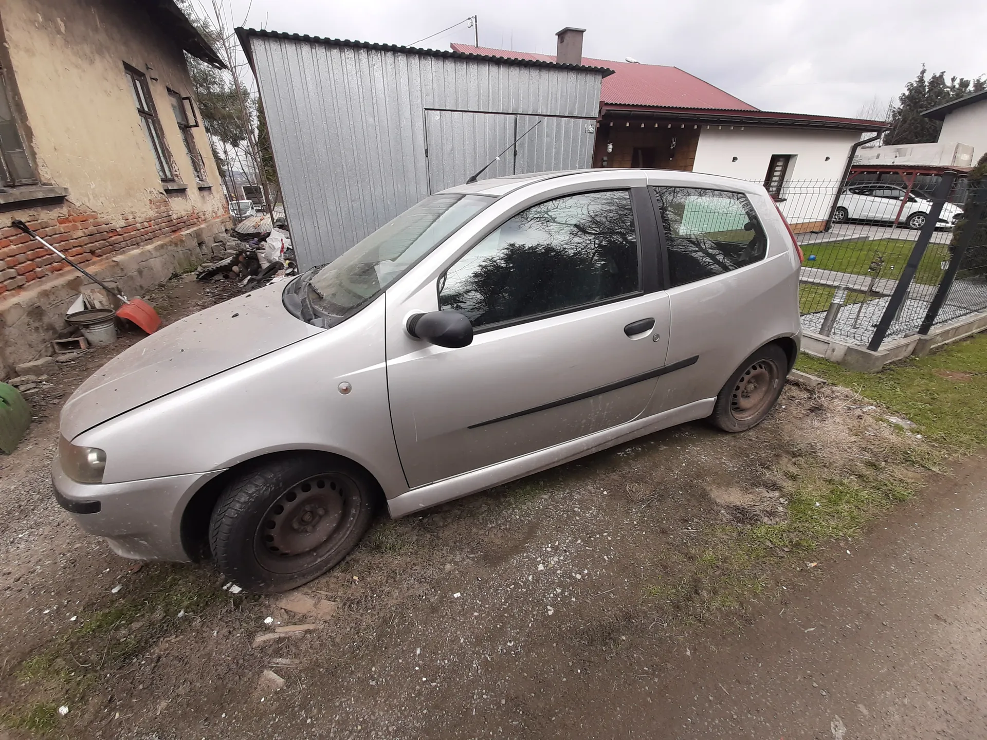 Srebrny samochód osobowy typu hatchback zaparkowany na podwórku z ziemią i żwirem. Po lewej stronie widoczny stary budynek z ceglanym fundamentem i uszkodzoną elewacją, obok metalowy garaż. W tle dom z czerwonym dachem, biała furtka oraz biały samochód za ogrodzeniem. Na podwórku znajdują się różne przedmioty, w tym wiadro i czerwony pojemnik. Niebo jest zachmurzone.