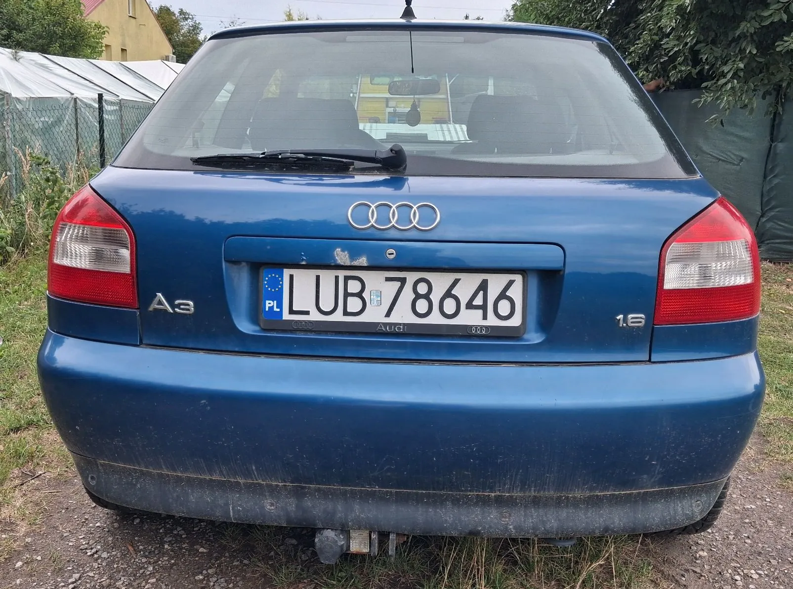 Tylny widok niebieskiego samochodu marki Audi A3 1.6. Na klapie bagażnika znajduje się logo Audi oraz tablica rejestracyjna o numerze LUB 78646. Samochód jest lekko zabrudzony w dolnej części zderzaka i posiada hak holowniczy.