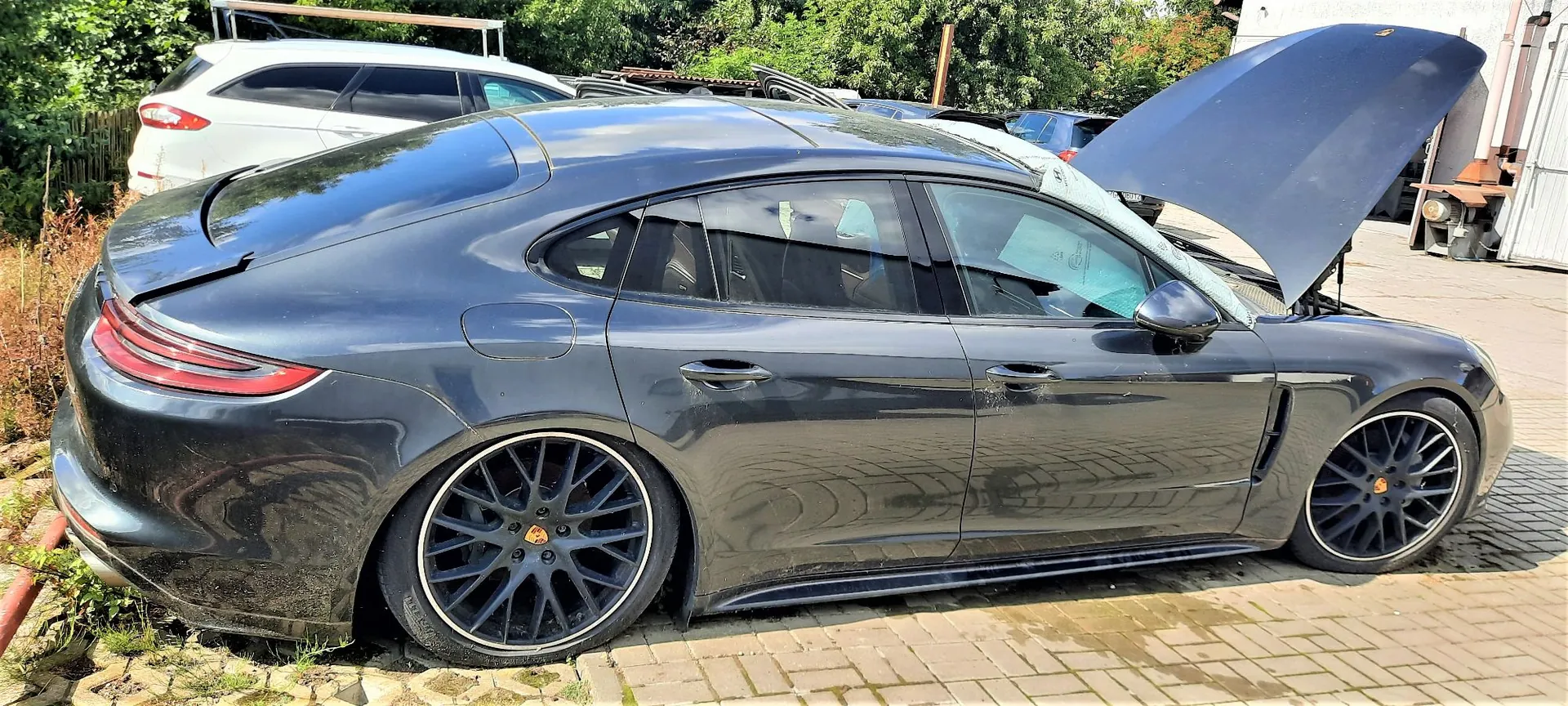 Profil ciemnoszarego samochodu marki Porsche Panamera zaparkowanego na kostce brukowej. Samochód ma otwartą maskę silnika i czarne felgi z logo marki. W tle widoczny jest biały samochód oraz zieleń drzew.