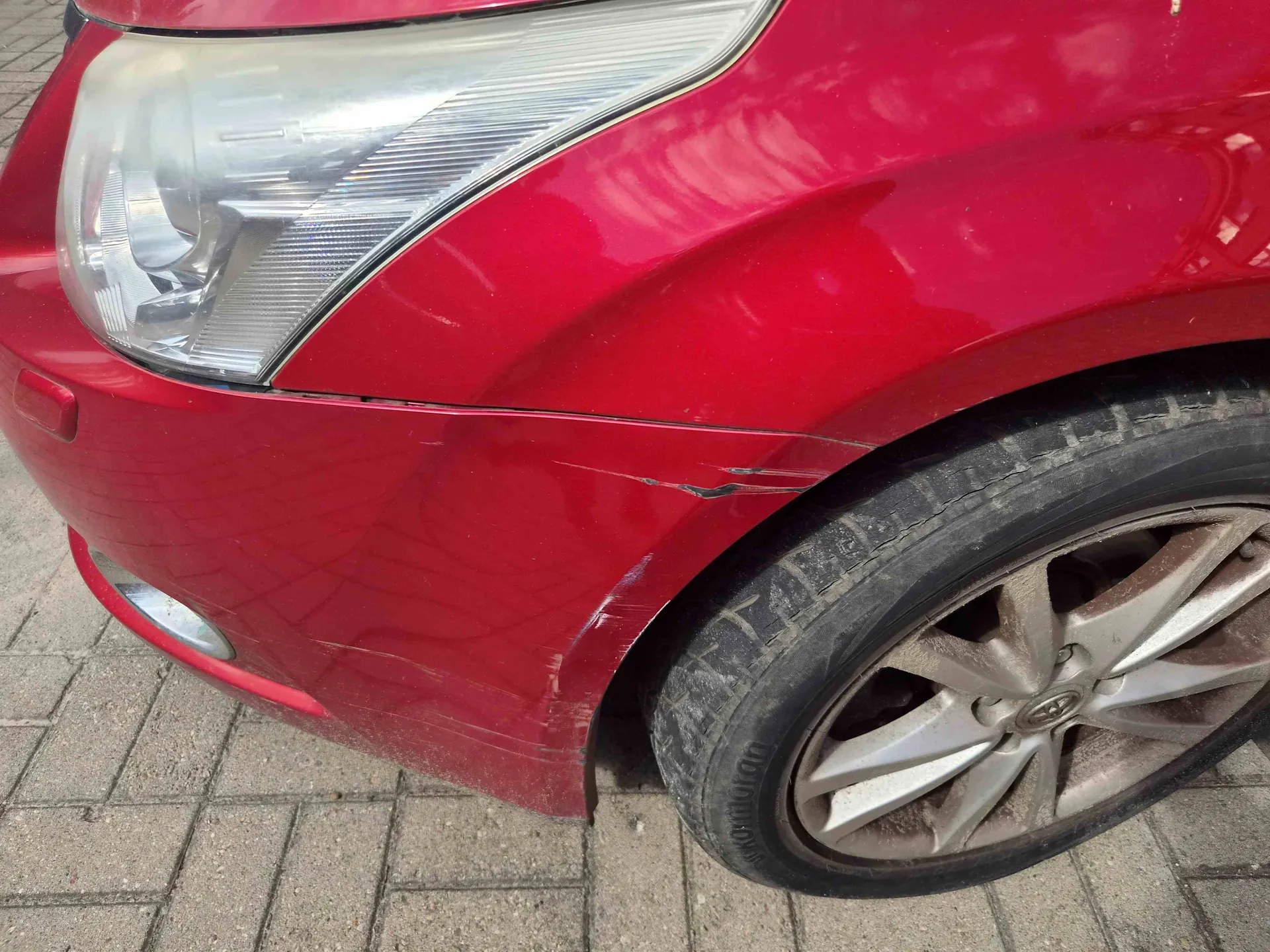 Zbliżenie na przedni prawy narożnik czerwonego samochodu. Widoczny jest reflektor, fragment maski oraz koło z aluminianą felgą. Na czerwonym zderzaku, nad nadkolem, znajdują się zarysowania i wgniecenia lakieru.