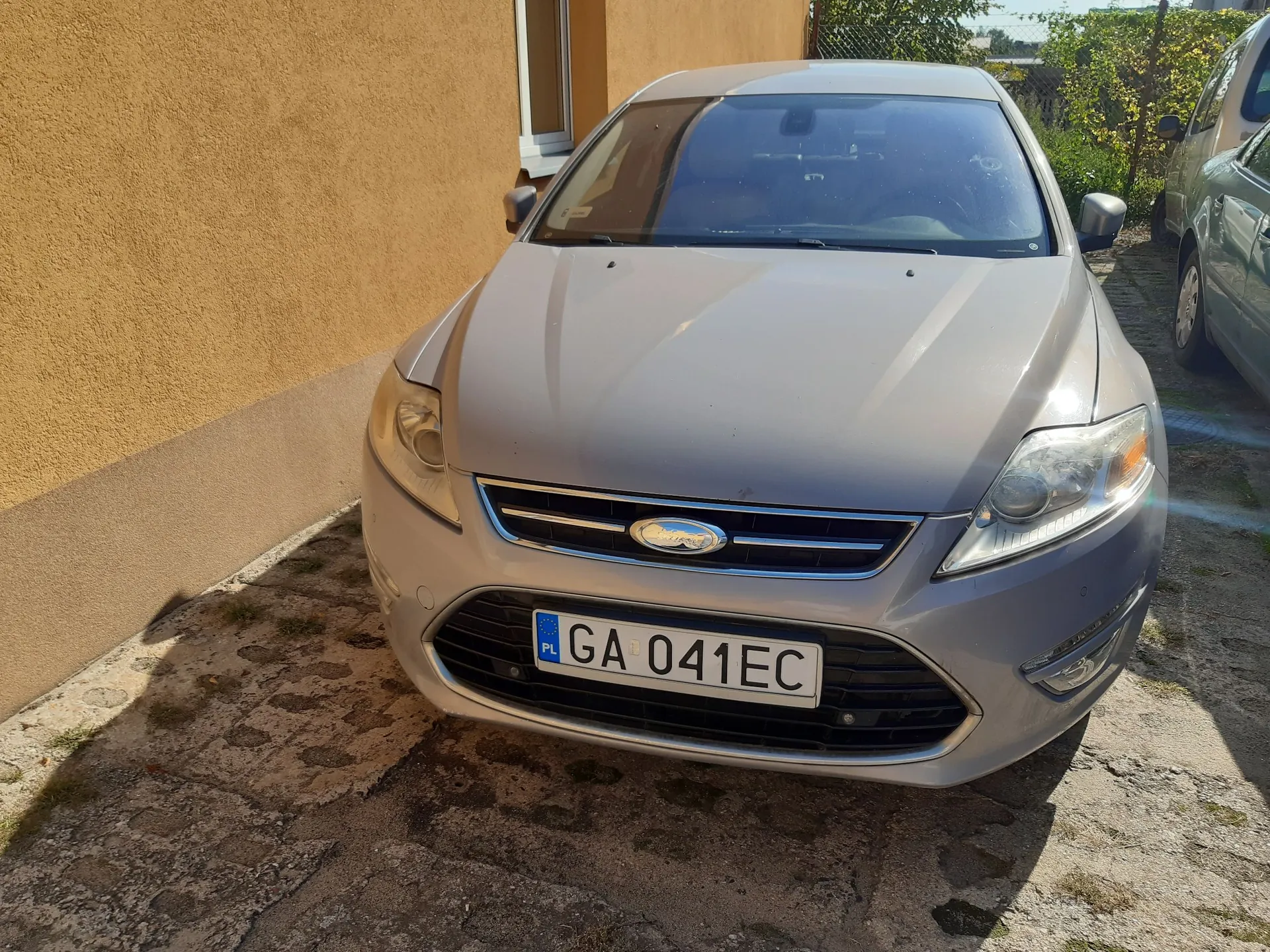 Srebrny samochód marki Ford zaparkowany na betonowym podjeździe obok żółtej ściany budynku. Widoczny jest przód pojazdu z tablicą rejestracyjną o numerze GA 041EC.