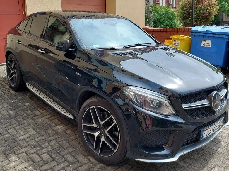 Czarny SUV marki Mercedes-Benz zaparkowany na brukowanej powierzchni przed budynkiem z czerwonymi drzwiami garażowymi. W tle widoczne są żółty i niebieski pojemnik na śmieci oraz zieleń. Tablica rejestracyjna: FZA 0650. Samochód posiada aluminiowe felgi i emblemat marki na przodzie.