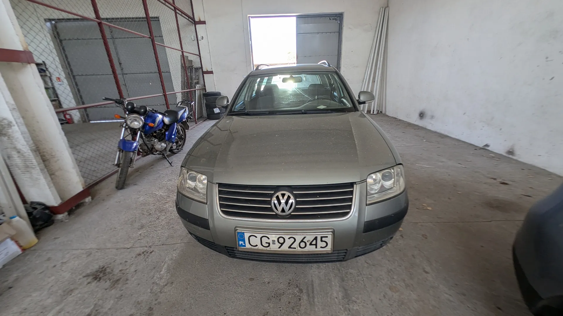Szary samochód Volkswagen z tablicą rejestracyjną CG-92645 (PL) stojący w garażu. Po lewej stronie niebieski motocykl, w tle siatka ochronna, stos opon i otwarte drzwi. Podłoga betonowa, białe ściany z widocznymi plamami.