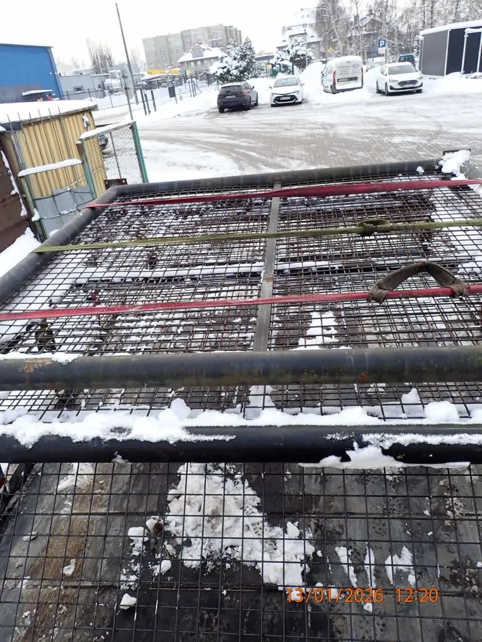 Zimowy krajobraz z metalowym ogrodzeniem pokrytym śniegiem. Ogrodzenie składa się z czarnych pionowych słupów i siatki. Na siatce widoczne są czerwone i zielone paski oraz brązowy uchwyt. Po lewej stronie obrazu znajduje się pomarańczowy znacznik czasu: 13/01/2026 12:20. W tle zasypana śniegiem droga z zaparkowanymi samochodami, budynki mieszkalne oraz struktura z niebieskim dachem. Śnieg pokrywa również powierzchnię ziemi i elementy ogrodzenia.