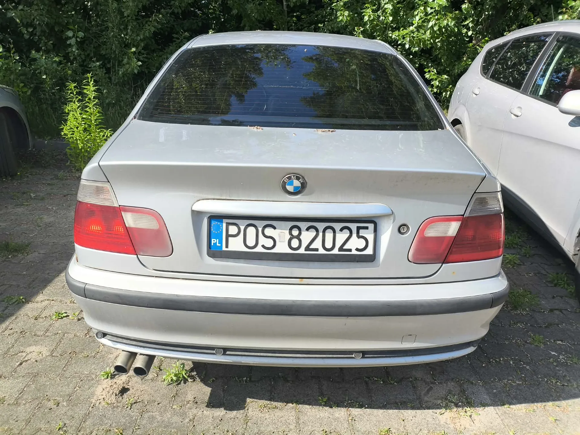 Tylny widok srebrnego samochodu marki BMW. Na klapie bagażnika znajduje się logo BMW oraz polska tablica rejestracyjna o numerze POS 82025. Samochód ma czerwone tylne lampy i podwójną rurę wydechową po lewej stronie. W tle widoczne są zielone krzewy i fragment innego, białego samochodu po prawej stronie.