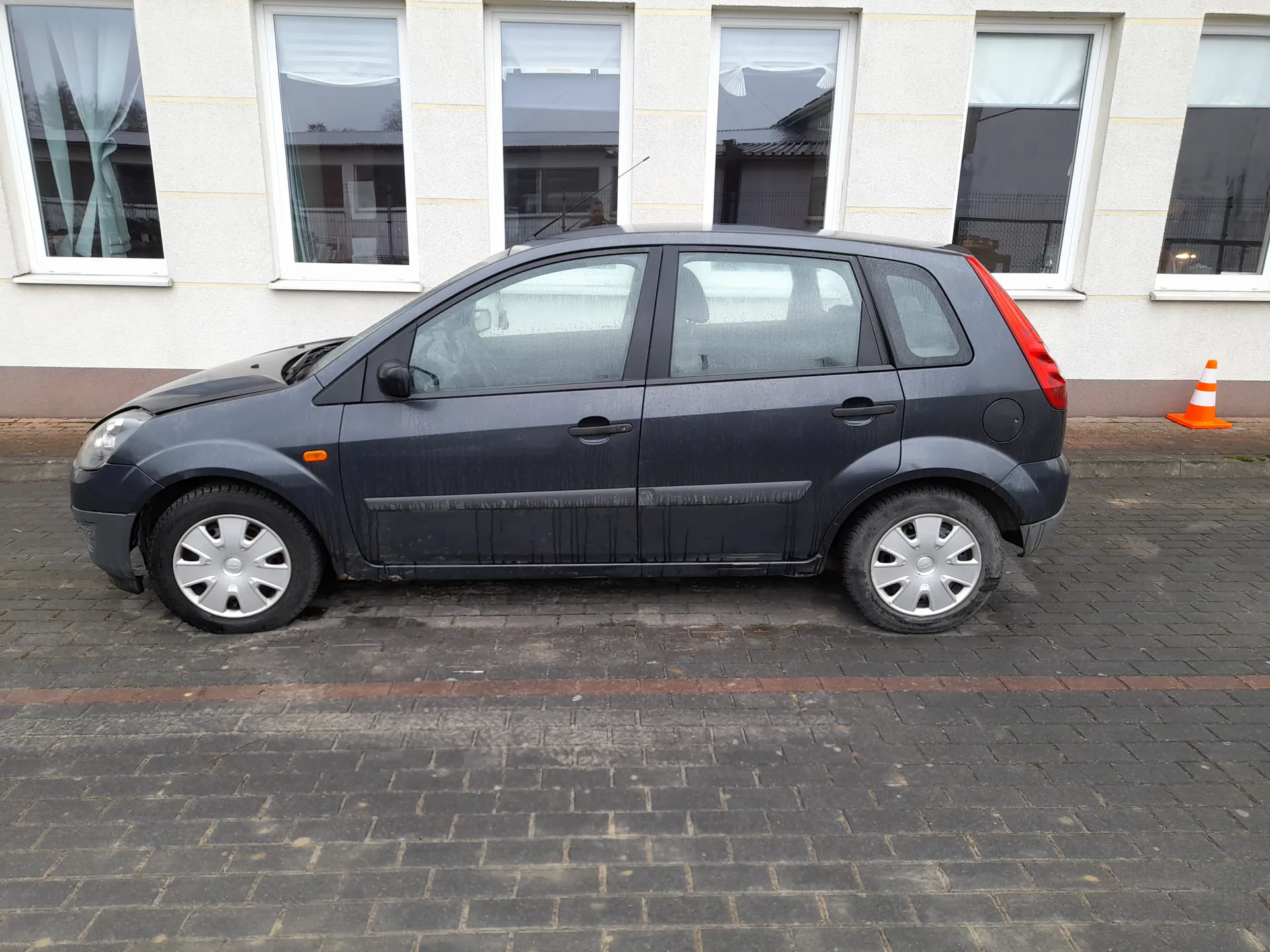 Ciemnoszary samochód osobowy typu hatchback zaparkowany na brukowanej ulicy. Pojazd jest zabrudzony, z widocznymi plamami błota na bokach. Samochód ma srebrne felgi i czerwone światło tylnego. W tle budynek z białymi ścianami i sześcioma oknami z białymi ramami, w niektórych oknach zasłony. Po prawej stronie obok budynku pomarańczowo-biały stożek drogowy. Ulica wyłożona jest ciemnymi kamieniami brukowymi.
