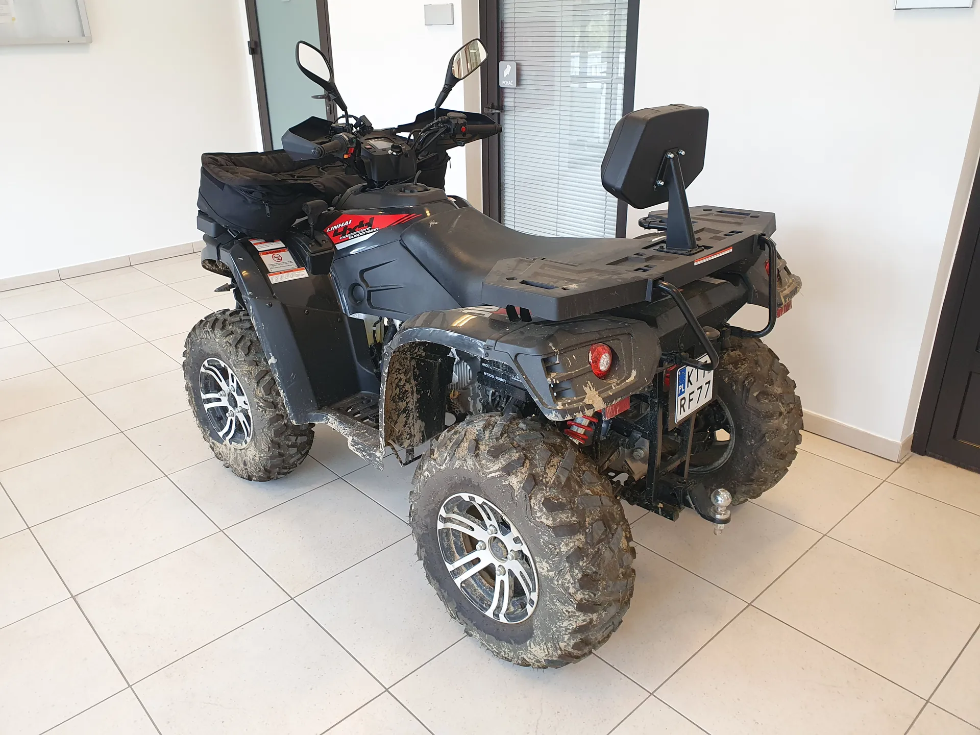 Czarny quad z dużymi oponami w błocie, stojący na białej płytkowej podłodze w pomieszczeniu. Widoczne elementy: czarne siedzenie, lusterka, tablica rejestracyjna PL K1 RF77, napisy 'LONCIN' i '400' na maszynie, czerwone światło tylnego reflektora, bagażnik z czarną torbą. W tle białe ściany, drzwi z przeszkleniem i roletą.