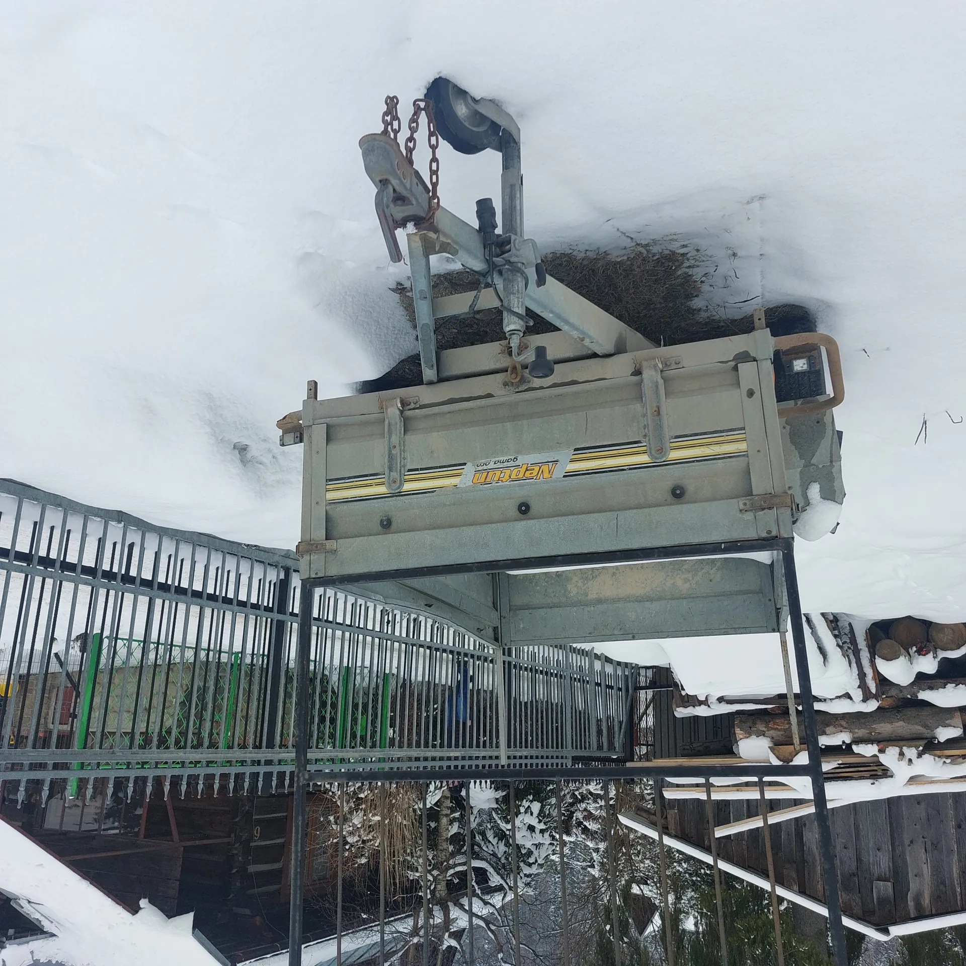 Przyczepa o szarym kolorze z żółtymi paskami i widocznym napisem 'Neptun' na boku, stojąca na śnieżnym terenie. W tle metalowe ogrodzenie, drewniany budynek z pokrytą śniegiem dachówką oraz drzewa i krzewy pokryte śniegiem. Na przyczepie widoczne są łańcuchy i elementy mechaniczne.