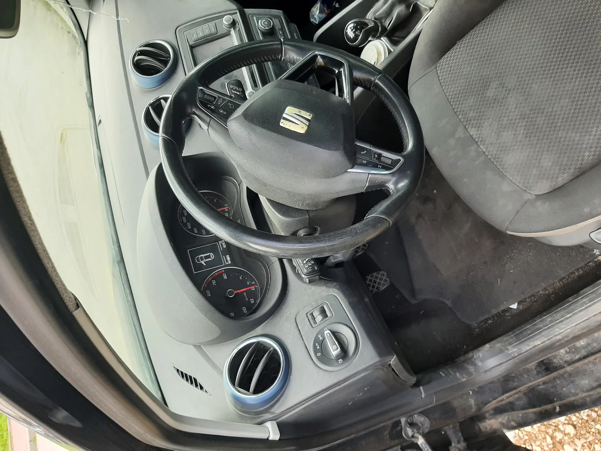 Wnętrze samochodu z kierownicą marki SEAT, deską rozdzielczą z licznikami prędkości, wentylatorami powietrza, przyciskami sterowania oraz częścią siedzenia pasażera. Widoczne są również fragmenty drzwi i podłogi z czarnym dywanikiem.