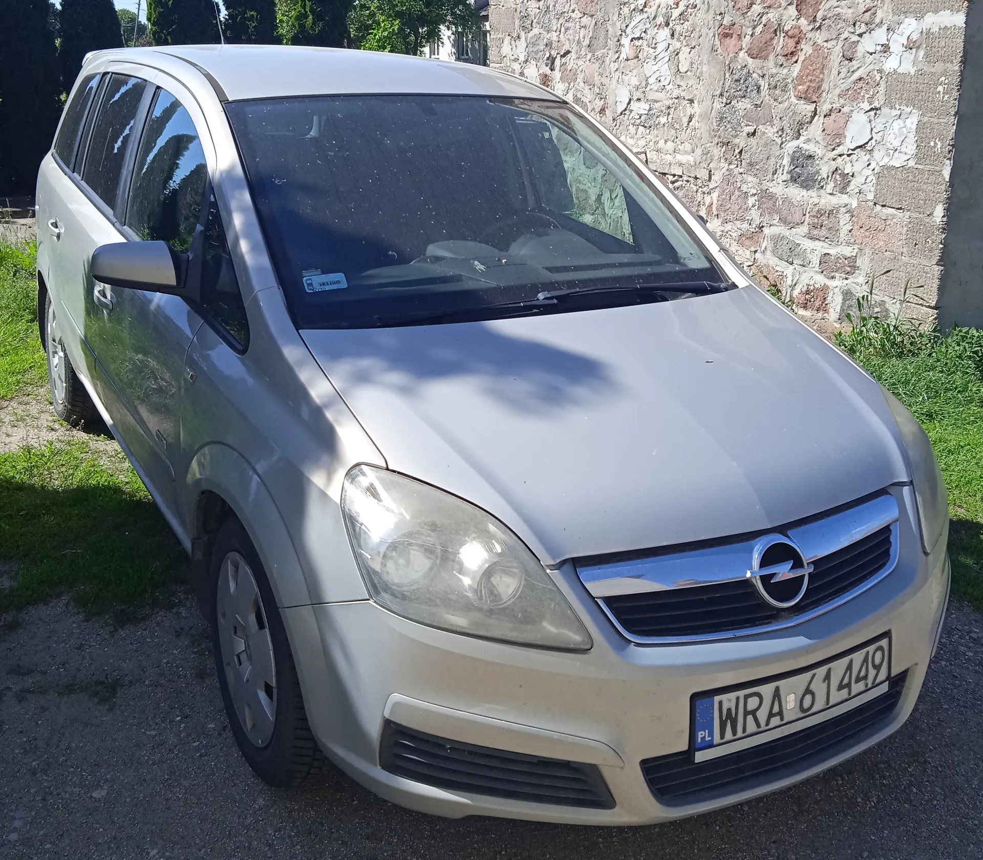 Srebrny samochód Opel zaparkowany na żwirowej powierzchni. Widoczna tablica rejestracyjna PL WRA 61449, logo Opel na masce, niebieski naklejka na przedniej szybie. Tło: kamienna ściana, zielona trawa i drzewa. Samochód ma pięć drzwi, reflektory i lusterka boczne.