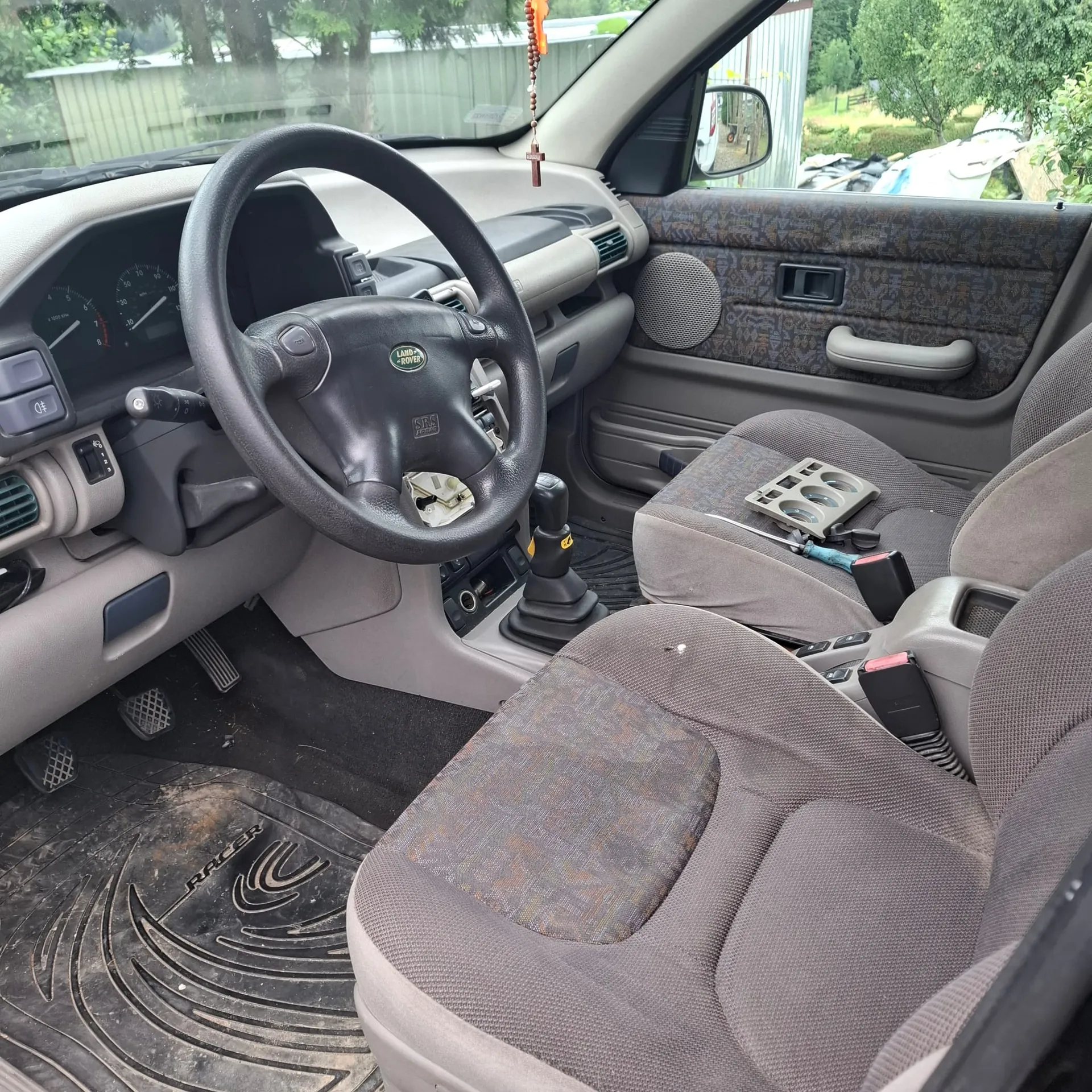 Wnętrze samochodu z perspektywy pasażera. Widoczna jest kierownica z logo Land Rover, deska rozdzielcza, dwa szare fotele z wzorzystą tapicerką oraz gumowa mata podłogowa z napisem RACER. Na siedzeniu pasażera leży mała taca z przegródkami i narzędzie.
