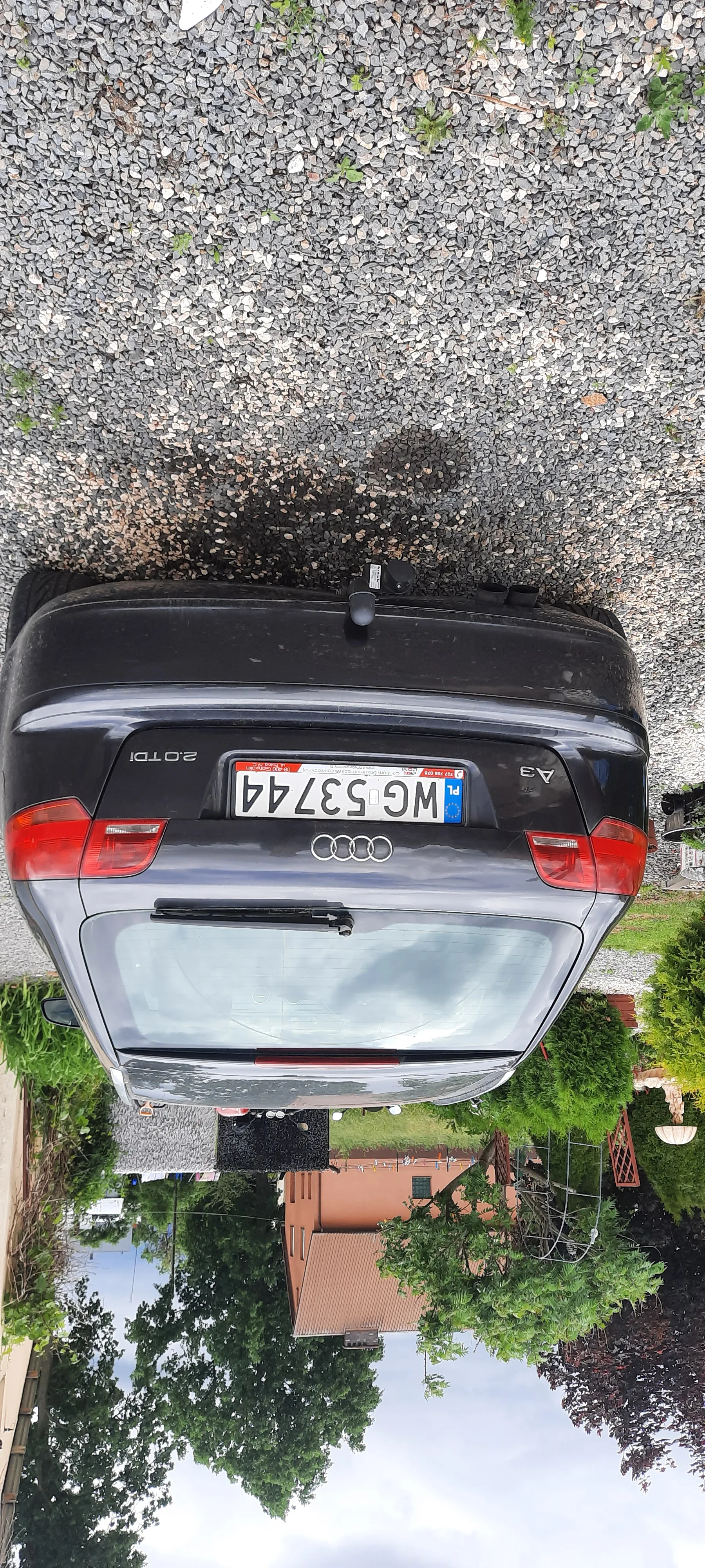 Ciemnoszary samochód Audi A3 2.0 TDI z tablicą rejestracyjną WG 53744 zaparkowany na podłożu z żwiru. Na tablicy rejestracyjnej widoczny niebieski pas z flagą UE oraz oznaczenie 'PL'. W tle dom z brązowym dachem, drzewa i krzewy.