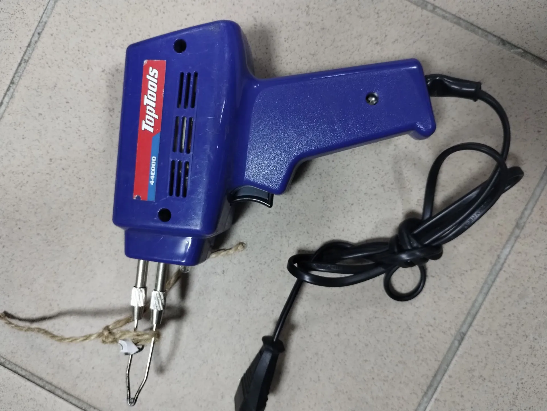 Niebieska lutownica elektryczna marki TopTools, model 44E000, z czarnym kablem zasilającym. Urządzenie leży na jasnych, ceramicznych płytkach. Na obudowie widoczne jest logo 'TopTools' oraz otwory wentylacyjne.