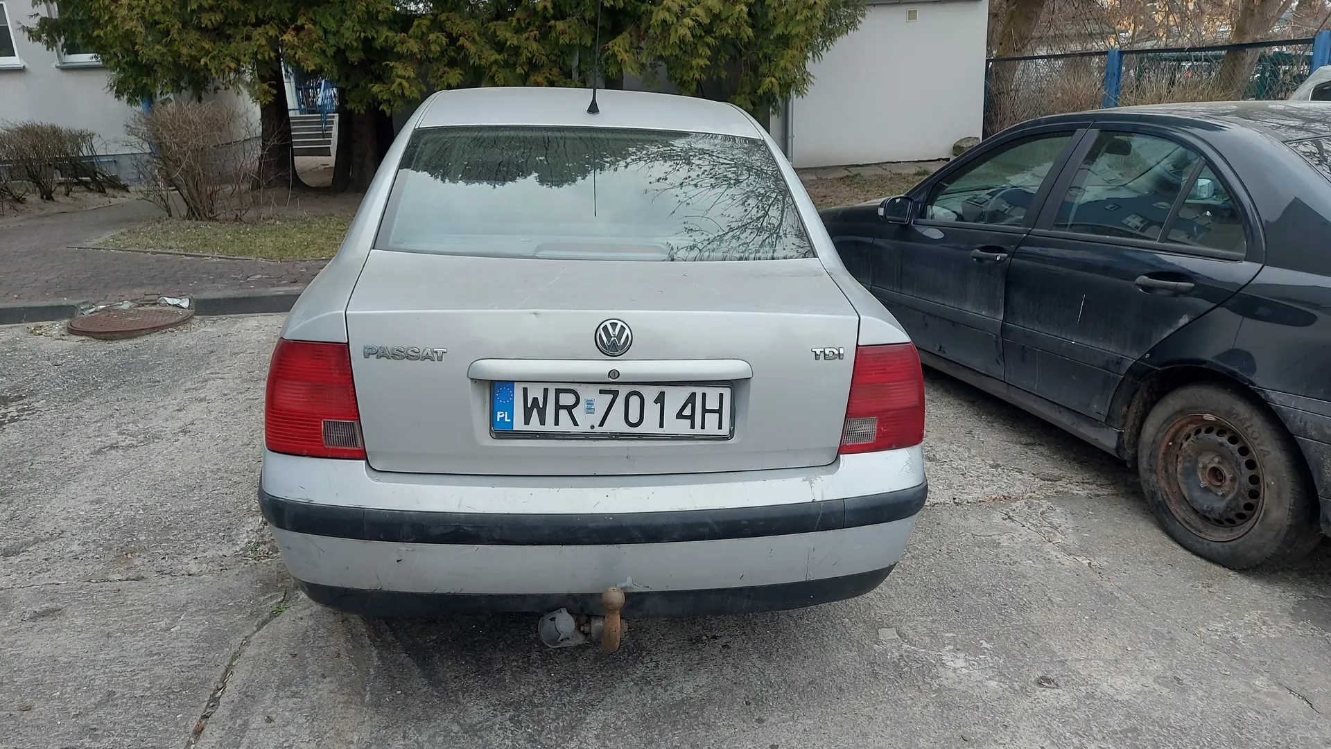 Z tyłu srebrny samochód Volkswagen Passat TDI z tablicą rejestracyjną WR 7014H (PL). Po prawej stronie ciemny, zabrudzony samochód osobowy z rdzawym kołem. Tło: drzewa, budynek z oknami, krzewy, ogrodzenie i betonowa powierzchnia z pęknięciami.