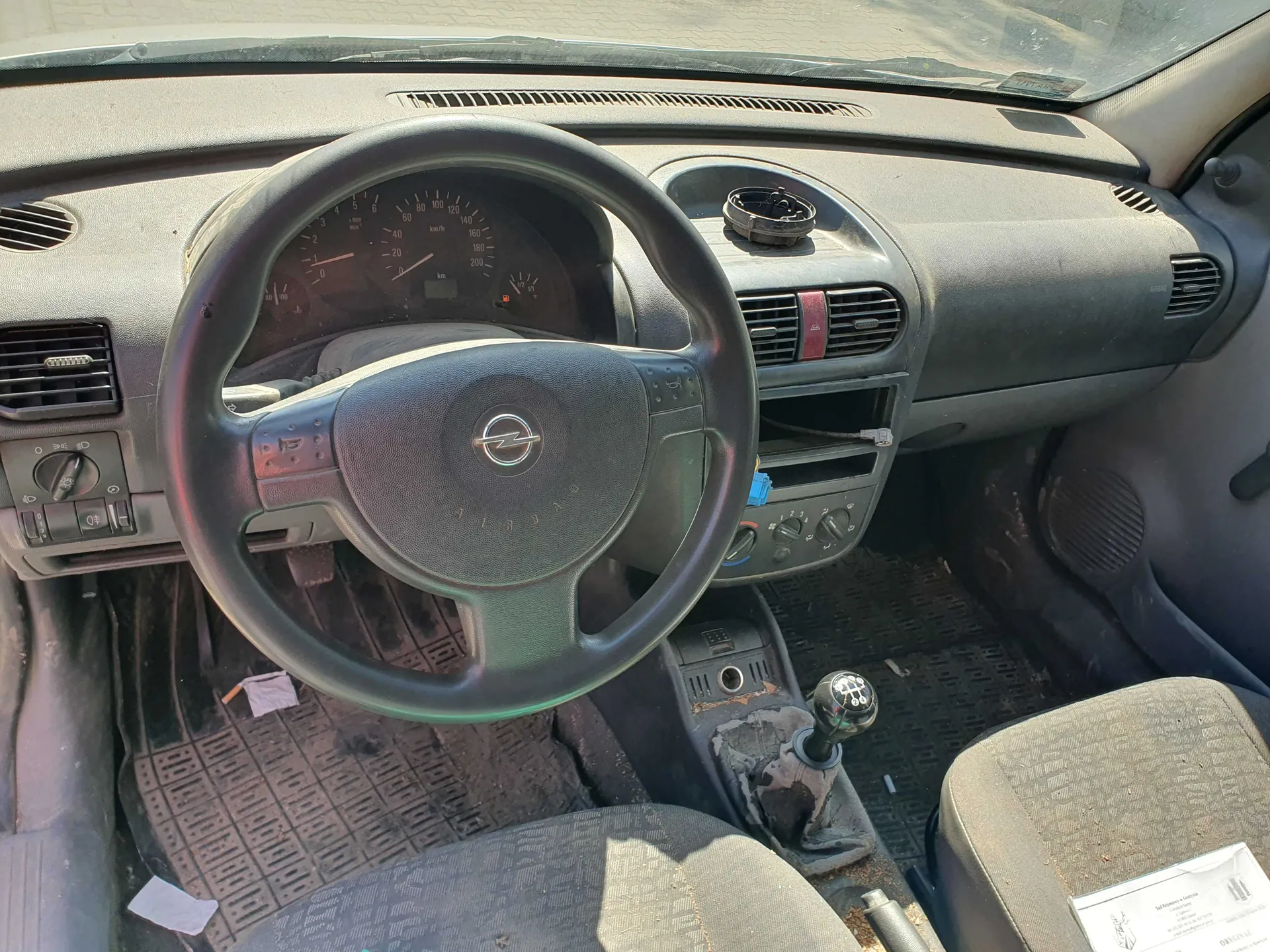 Wnętrze samochodu marki Opel, widok z perspektywy kierowcy. Widoczna jest czarna kierownica z logo producenta w centrum, deska rozdzielcza z analogowymi wskaźnikami prędkości i obrotów, otwory nawiewów oraz konsola środkowa z lewarkiem zmiany biegów. Wnętrze jest zakurzone, a na podłodze znajdują się gumowe dywaniki.