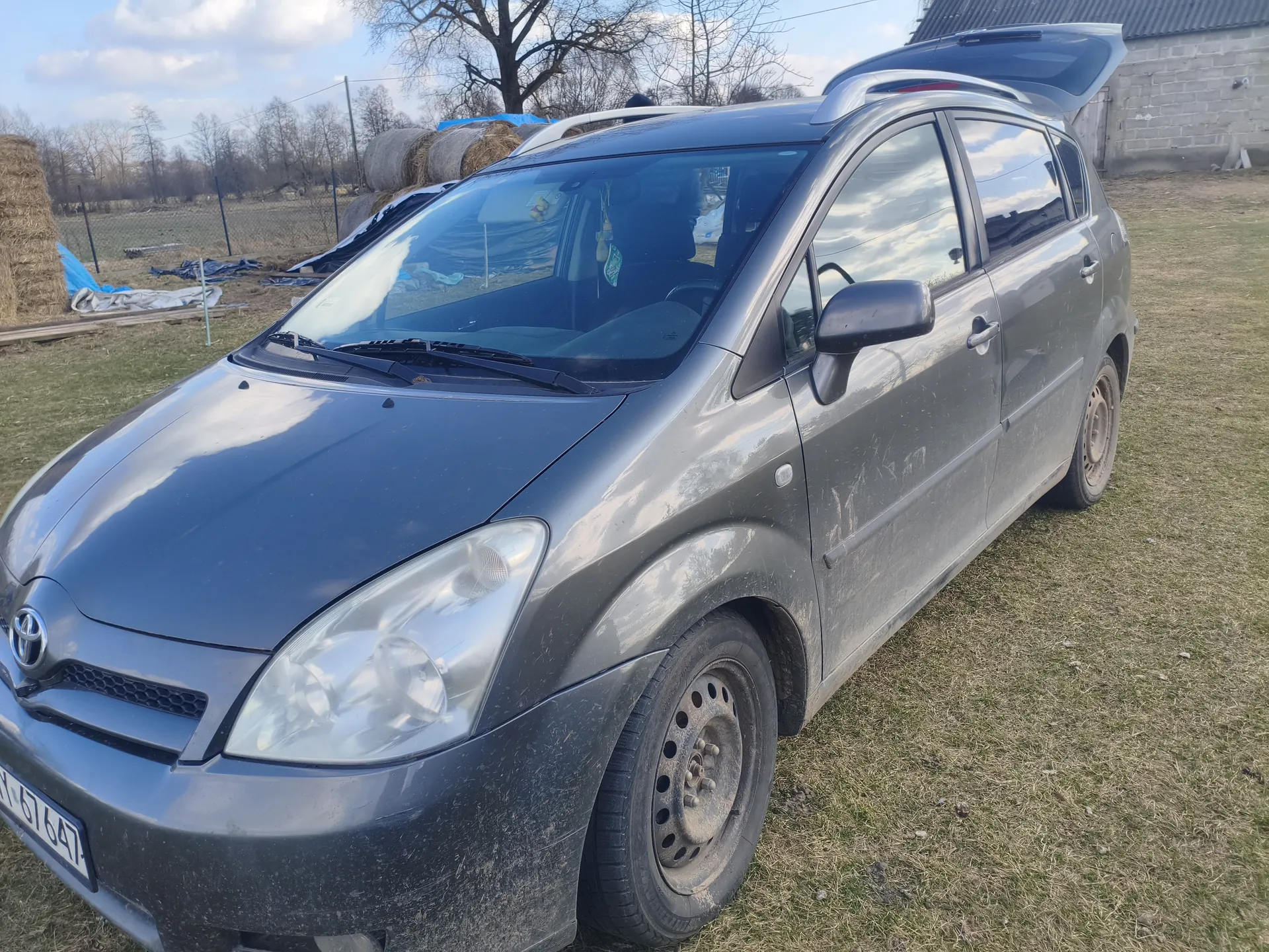 Szary samochód osobowy marki Toyota z otwartym bagażnikiem, zaparkowany na trawie. Widoczna tablica rejestracyjna N 67647. Samochód jest zabrudzony, na tle sterty siana, ogrodzenia, drzew bez liści oraz betonowego budynku. Niebo częściowo zachmurzone.