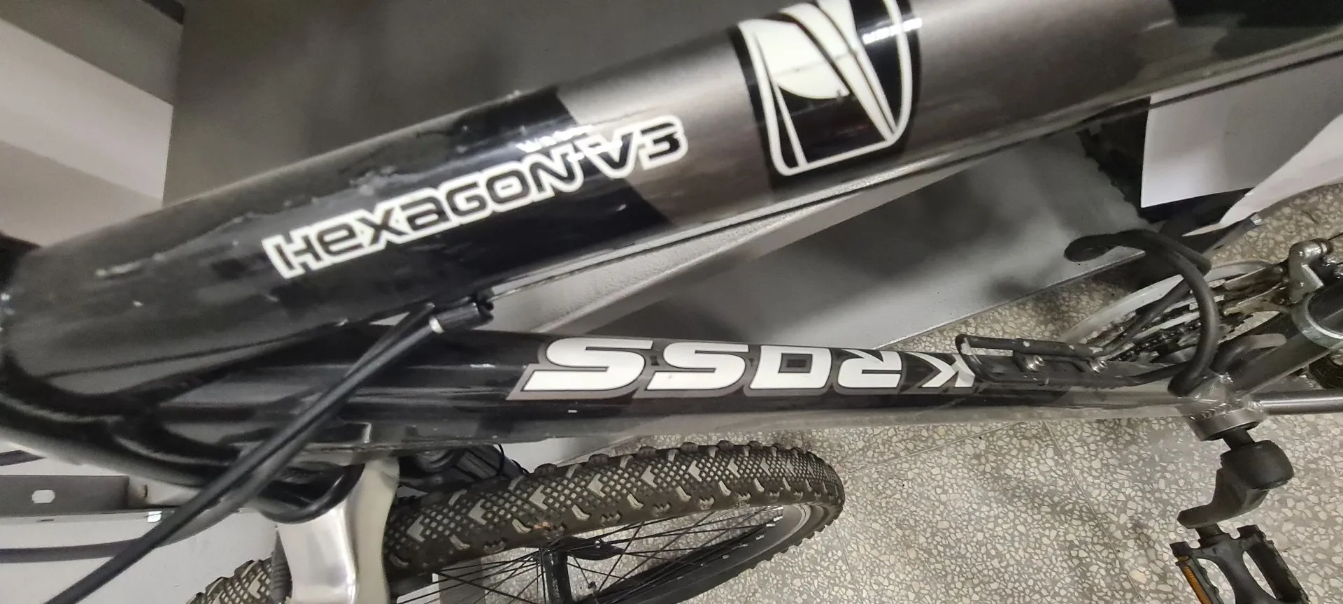Zbliżenie na czarną ramę roweru z białymi napisami. Na górnej rurze widnieje napis „HEXAGON V3”, a na dolnej rurze napis „KROSS”. W kadrze widoczny jest fragment koła z terenową oponą oraz elementy układu hamulcowego i napędowego.