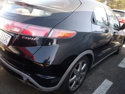 Czarny samochód Honda Civic widziany od tyłu, z widocznym numerem rejestracyjnym 915C oraz napisem CIVIC na bagażniku. Pojazd zaparkowany na asfaltowej powierzchni z białymi liniami parkingu. W tle częściowo widoczny inny samochód oraz drzewa.