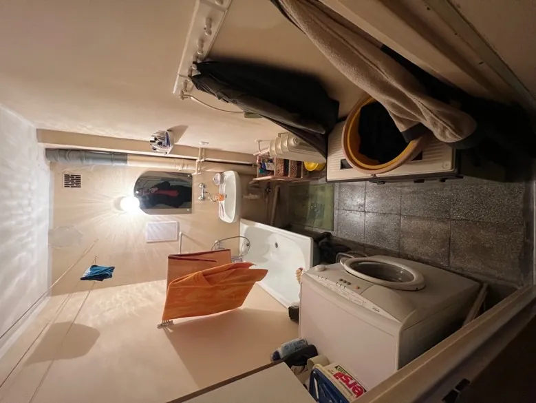 Wąska pralnia lub łazienka z beżowymi ścianami i podłogą wyłożoną szarymi płytkami. Po lewej stronie biała pralka, obok niej biała wanna z pomarańczowym ręcznikiem wiszącym na wieszaku. W głębi umywalka z lustrem, w którym odbija się osoba robiąca zdjęcie. Na umywalce i półkach znajdują się różne przedmioty. Po prawej stronie wieszaki z ciemnymi i jasnymi ubraniami, żółty kosz oraz zielony dywanik na podłodze. Na ścianie wiszą niebieski kawałek tkaniny i pionowa rura. Sufit oświetlony jest przez żarówkę.