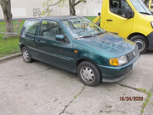 Zielony samochód osobowy typu hatchback zaparkowany na betonowej powierzchni. Obok niego znajduje się żółty samochód dostawczy. W tle widać białą ścianę z graffiti '88' i 'JFVF', drzewa oraz wózek zakupowy na trawniku. Na samochodzie zielonym widoczna tablica rejestracyjna. W prawym dolnym rogu obrazu umieszczona data '15/04/2026'.