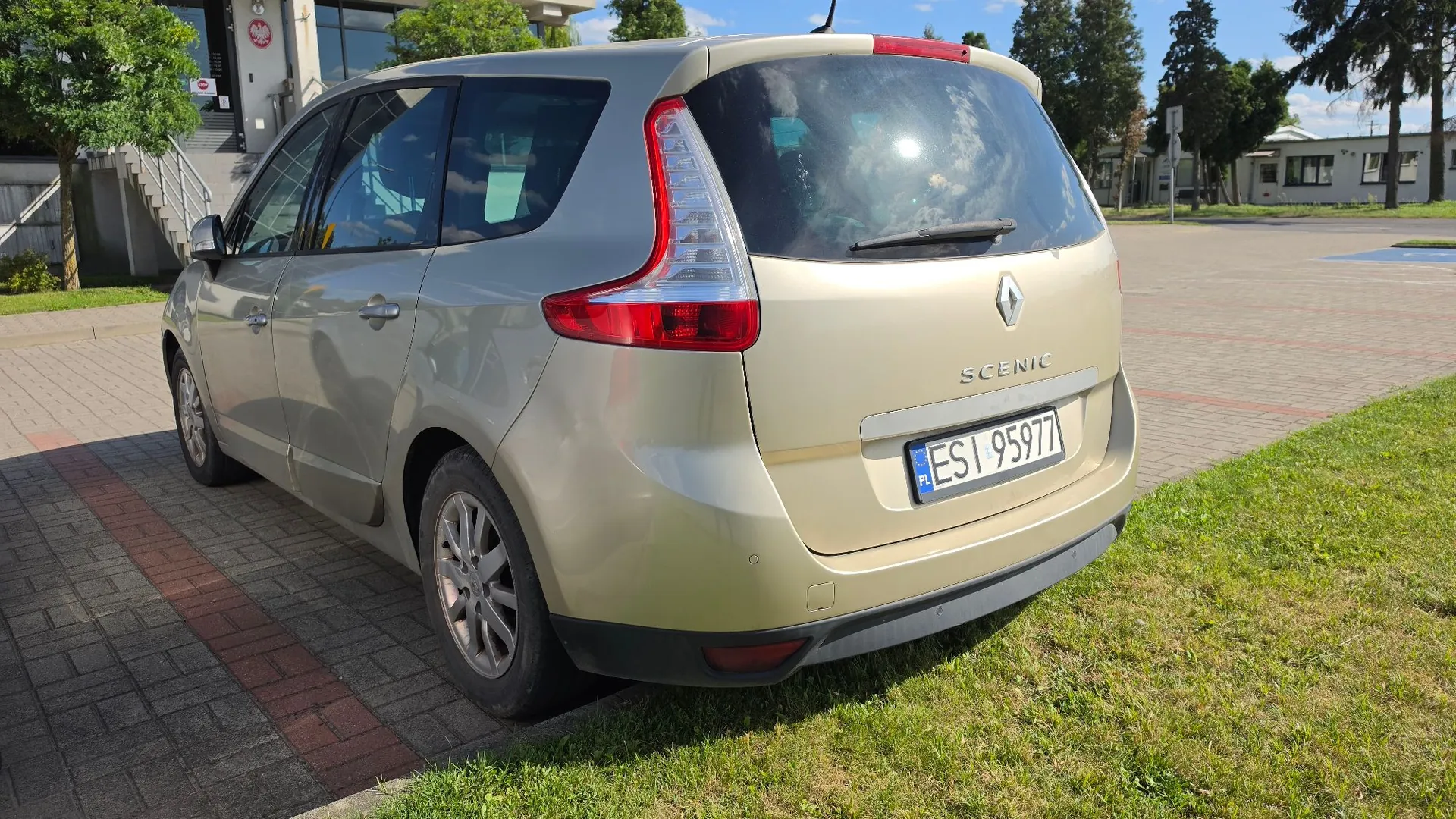Złoty samochód marki Renault Scenic zaparkowany na kostce brukowej obok trawnika. Widok z tyłu i z boku pojazdu. Samochód posiada polską tablicę rejestracyjną o numerze ESI 95977. W tle widoczne są budynki, drzewa i słoneczne niebo.