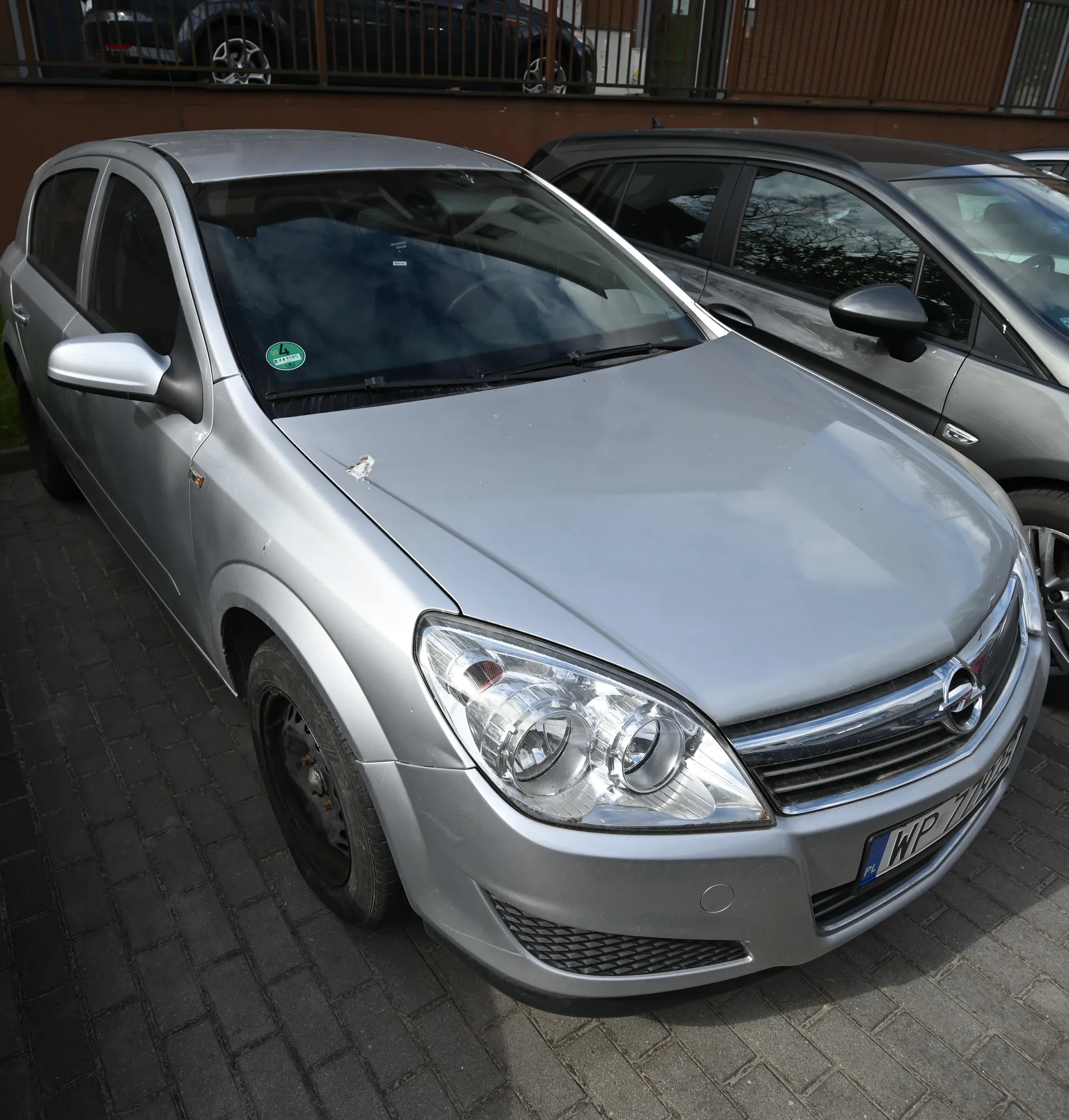 Srebrny samochód marki Opel zaparkowany na brukowanej powierzchni. Widoczna tablica rejestracyjna z napisem WP 719ES oraz zielony naklejka na przedniej szybie. W tle drugi samochód, ogrodzenie i fragment budynku. Samochód ma uszkodzenie na masce.