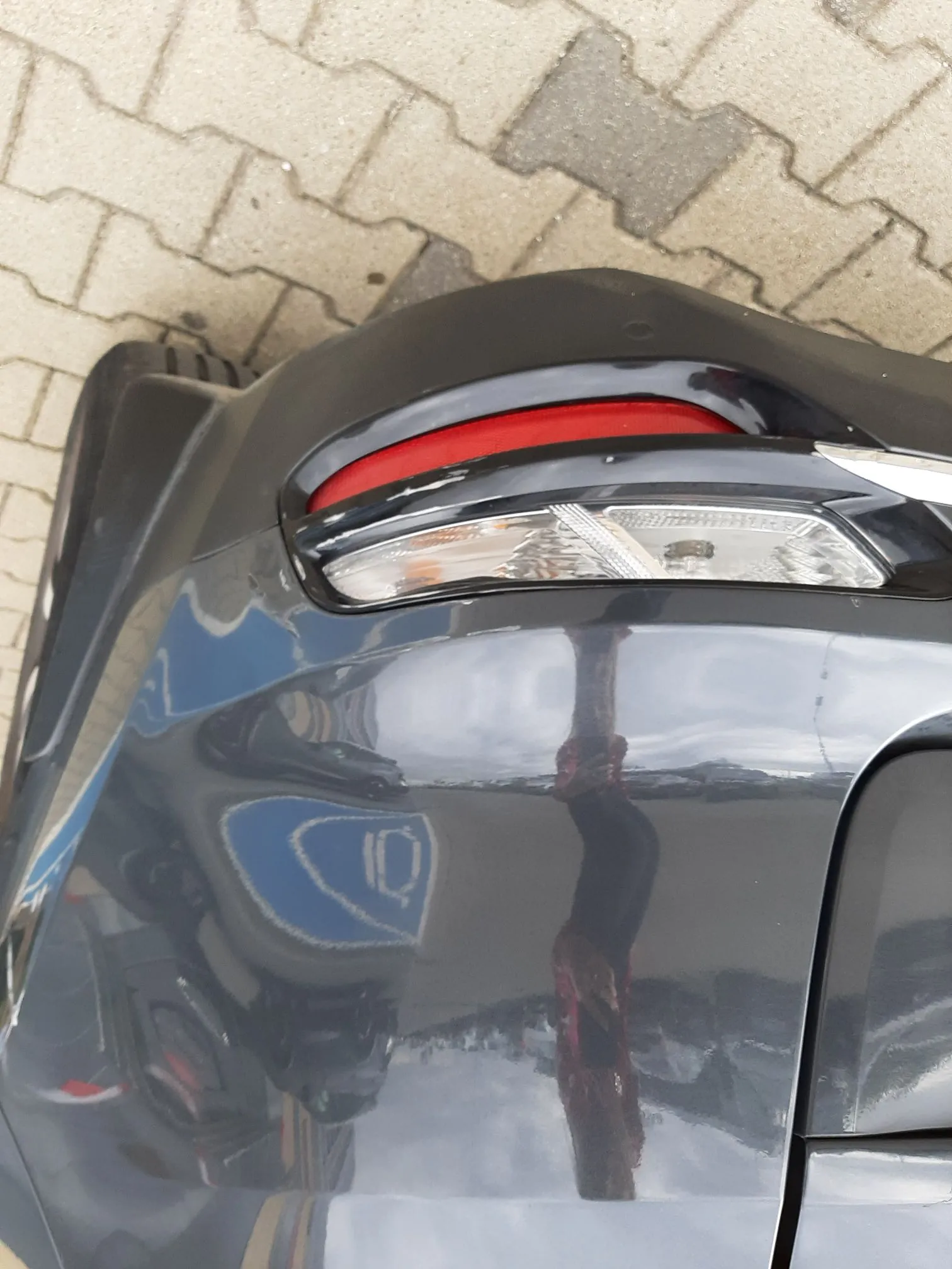 Zbliżenie na tylną część ciemnoszarego, błyszczącego samochodu. Widoczny jest fragment tylnej lampy z czerwonym i białym elementem oraz odblaski na karoserii. Samochód stoi na kostce brukowej.