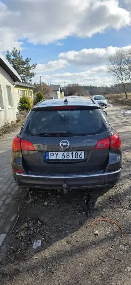 Z tyłu widoczny ciemnoszary samochód marki Opel z tablicą rejestracyjną PY 68186. Pojazd stoi na nierównym podłożu z drobnymi śmieciem i gałęziami. W tle dom z żółtymi ścianami, drzewa bez liści, droga oraz niebo z białymi chmurami. Drugi samochód widoczny w oddali na drodze.