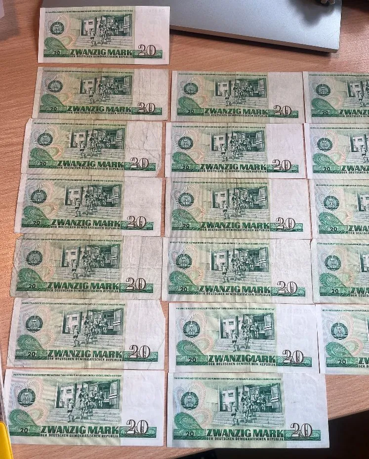 Zdjęcie przedstawiające kolekcję banknotów o nominale 20 marek z Niemieckiej Republiki Demokratycznej (NRD). Banknoty są ułożone w rzędach na brązowej powierzchni. Na każdym z nich widnieje napis „ZWANZIG MARK” oraz liczba „20”. Większość banknotów ma zielono-szarą kolorystykę z grafiką przedstawiającą budynki i postacie.