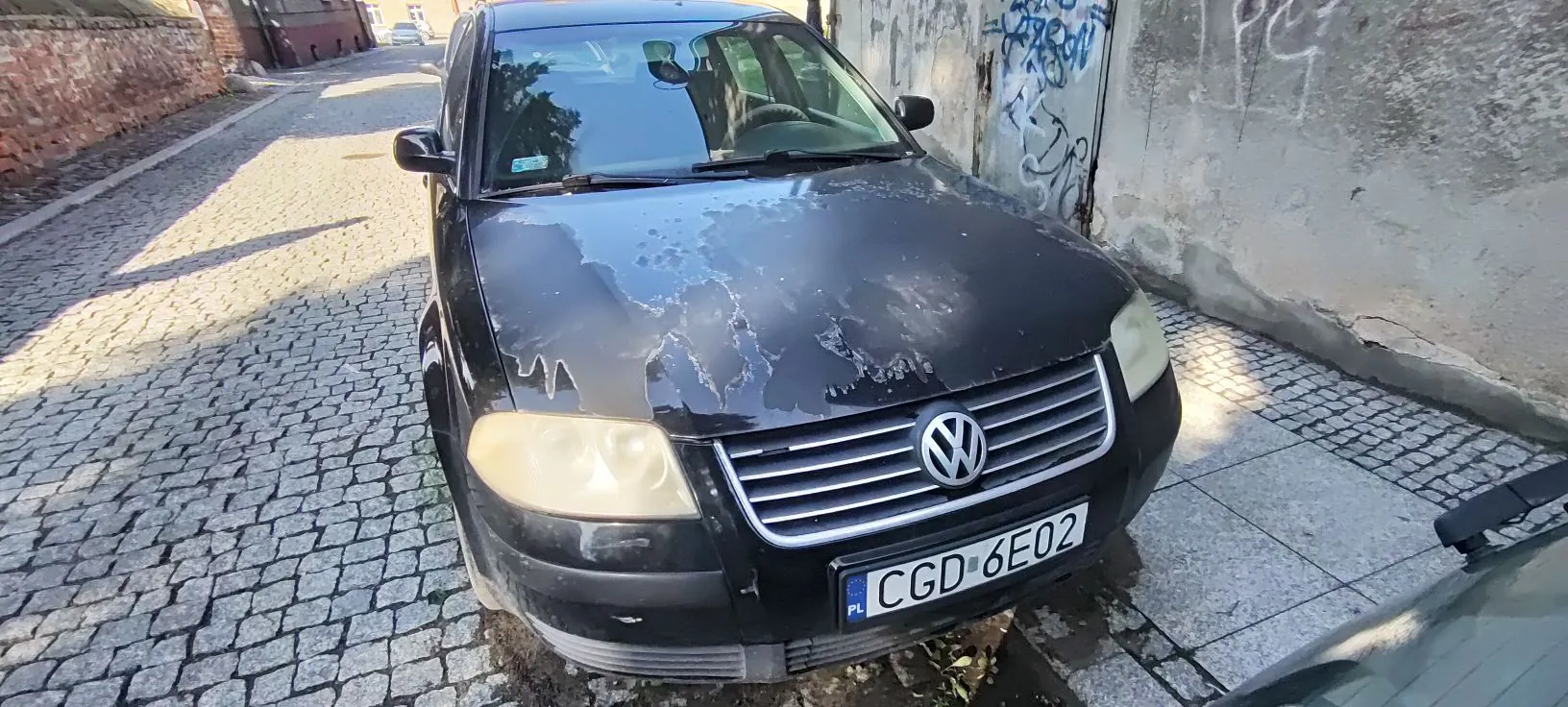 Czarny samochód marki Volkswagen zaparkowany na brukowanej uliczce obok jasnej, lekko zniszczonej ściany z graffiti. Lakier na masce samochodu jest mocno zniszczony i łuszczy się. Pojazd posiada polską tablicę rejestracyjną o numerze CGD 6E02.