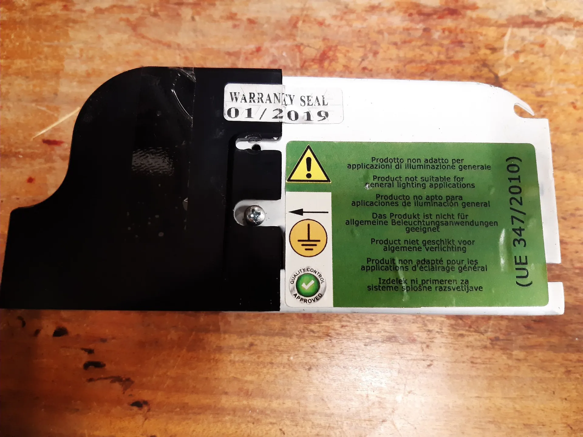 Zdjęcie białej etykiety technicznej przyklejonej do drewnianej powierzchni. W górnej części znajduje się naklejka z napisem „WARRANTY SEAL 01/2019”. Po prawej stronie widoczna jest zielona tabliczka ostrzegawcza z żółtym trójkątem z wykrzyknikiem, symbolem uziemienia oraz tekstem w kilku językach informującym, że produkt nie jest odpowiedni do ogólnych zastosowań oświetleniowych. Po prawej krawędzi widnieje pionowy napis „(UE 347/2010)”. W lewym dolnym rogu zielonej tabliczki znajduje się okrągły znak „QUALITY CONTROL APPROVED”. Lewa część etykiety jest zakryta czarną taśmą.
