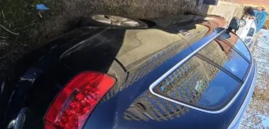 Czarny samochód osobowy zaparkowany na podłożu z żwiru i trawy. Widok od tyłu z czerwonymi światłami tylnymi. W tle widoczny inny pojazd oraz drzewa.