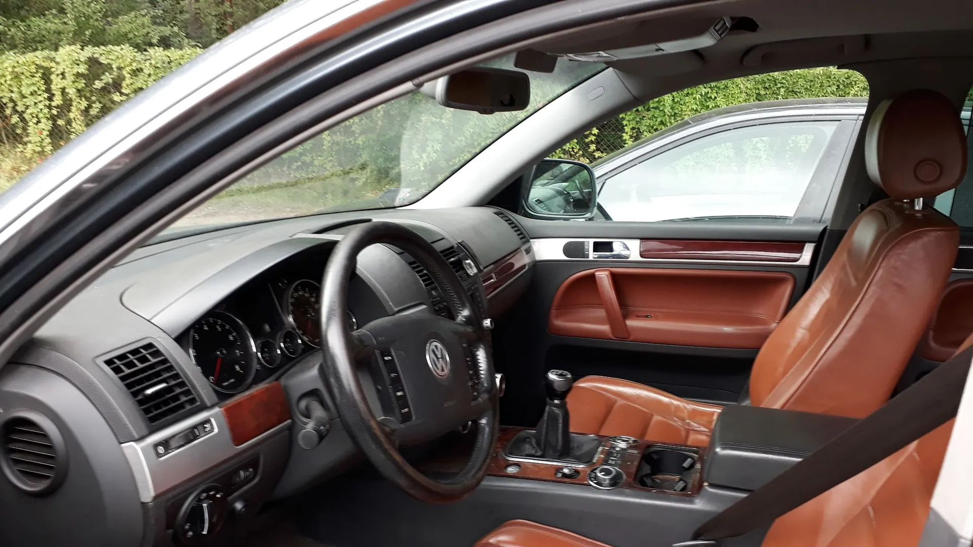 Wnętrze samochodu marki Volkswagen z widokiem na deskę rozdzielczą i fotele. Samochód posiada brązową, skórzaną tapicerkę na siedzeniach i panelach drzwi oraz ciemnoszarą deskę rozdzielczą z drewnianymi wstawkami. Widoczna jest kierownica z logo Volkswagena, zegary analogowe oraz lewarek zmiany biegów. Zdjęcie wykonane z perspektywy otwartych drzwi kierowcy.