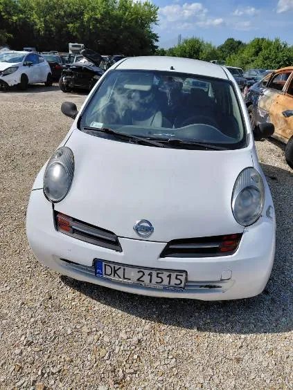 Widok z przodu białego samochodu marki Nissan zaparkowanego na żwirowym placu. Pojazd ma polską tablicę rejestracyjną o numerze DKL 21515. W tle widoczne są inne samochody oraz zielone drzewa pod jasnym niebem.