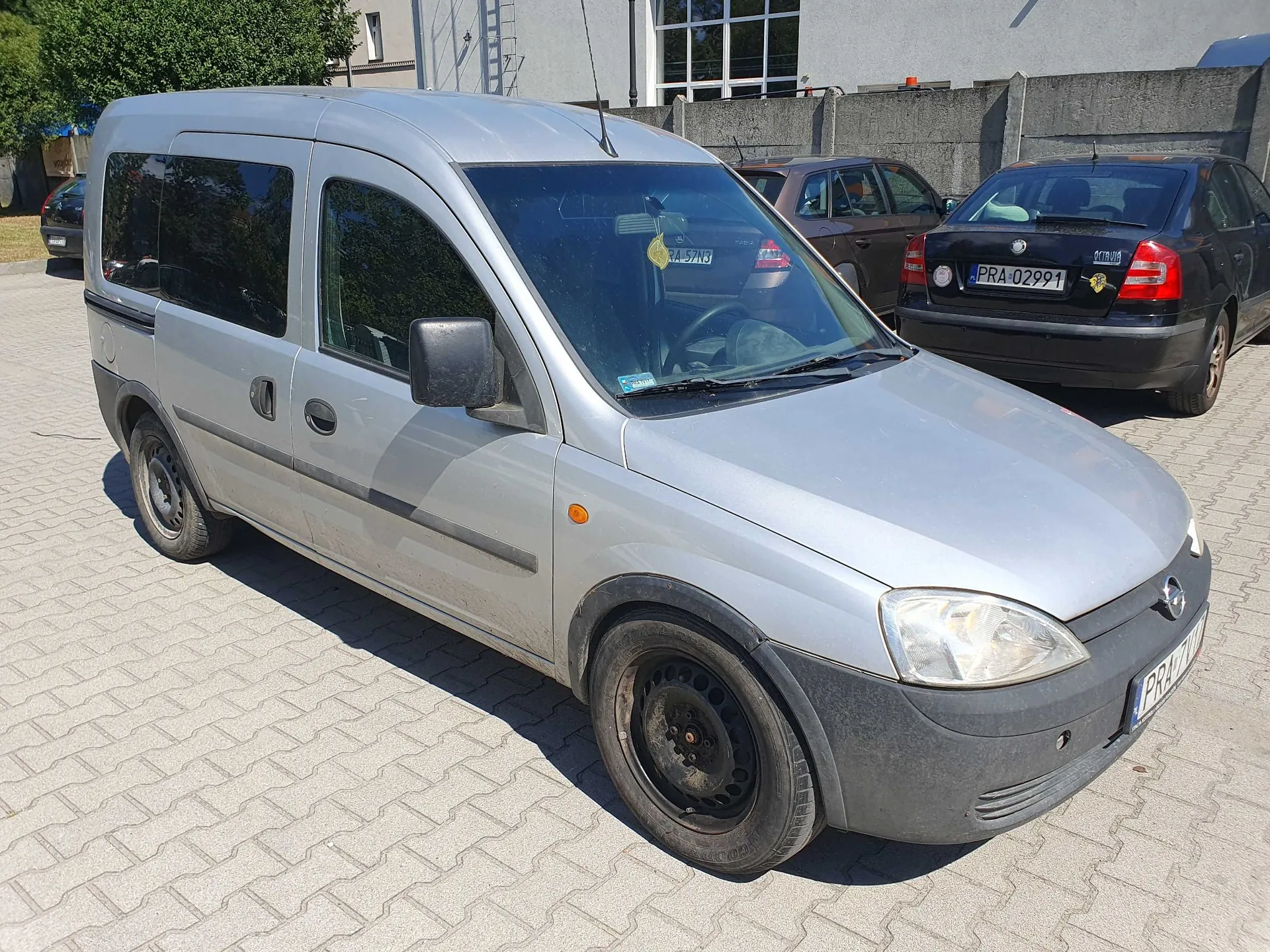 Srebrny samochód dostawczy marki Opel Combo zaparkowany na kostce brukowej. Pojazd jest widoczny z przodu i z boku. W tle znajdują się dwa inne samochody: brązowy i czarny, a także szary betonowy mur i fragment budynku.
