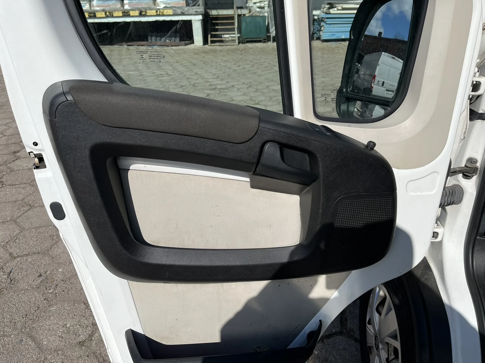 Zdjęcie wnętrza otwartych drzwi białego samochodu. Widoczny jest czarny plastikowy panel boczny z wbudowaną klamką i szarym elementem wykończeniowym w centralnej części. W górnej części znajduje się podłokietnik. Przez szybę widoczny jest plac z materiałami budowlanymi, a w lusterku bocznym odbija się biały samochód dostawczy.