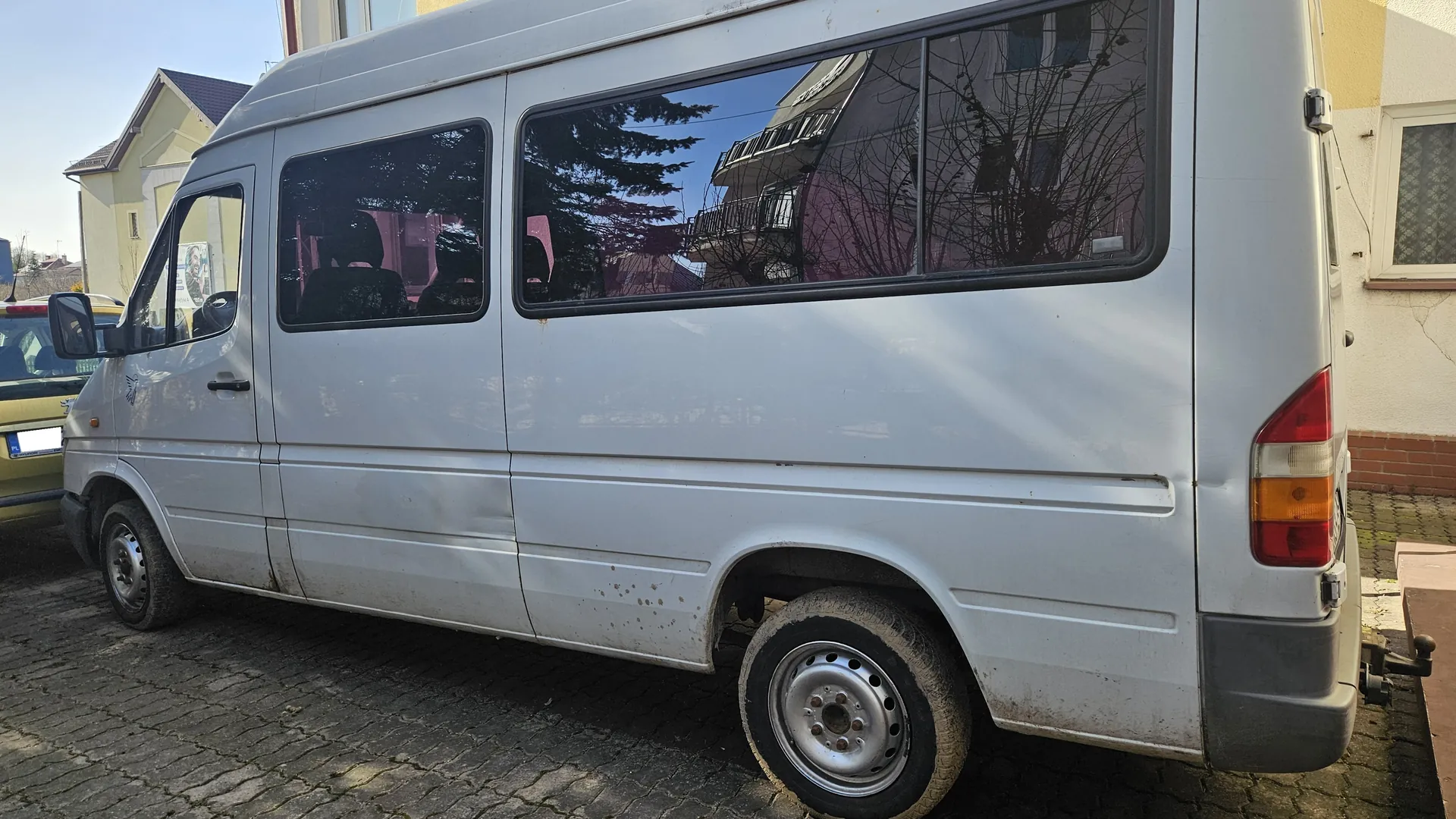 Srebrny furgon zaparkowany na brukowanej ulicy. Widoczna boczna i tylna część pojazdu z czterema oknami, odbijającymi drzewa i budynki. Furgon ma brudne plamy na karoserii i koła z metalowymi felgami. W tle częściowo widoczny żółty samochód z tablicą rejestracyjną oraz budynki mieszkalne z żółtymi i białymi elewacjami. Niebo jest jasne i bezchmurne.